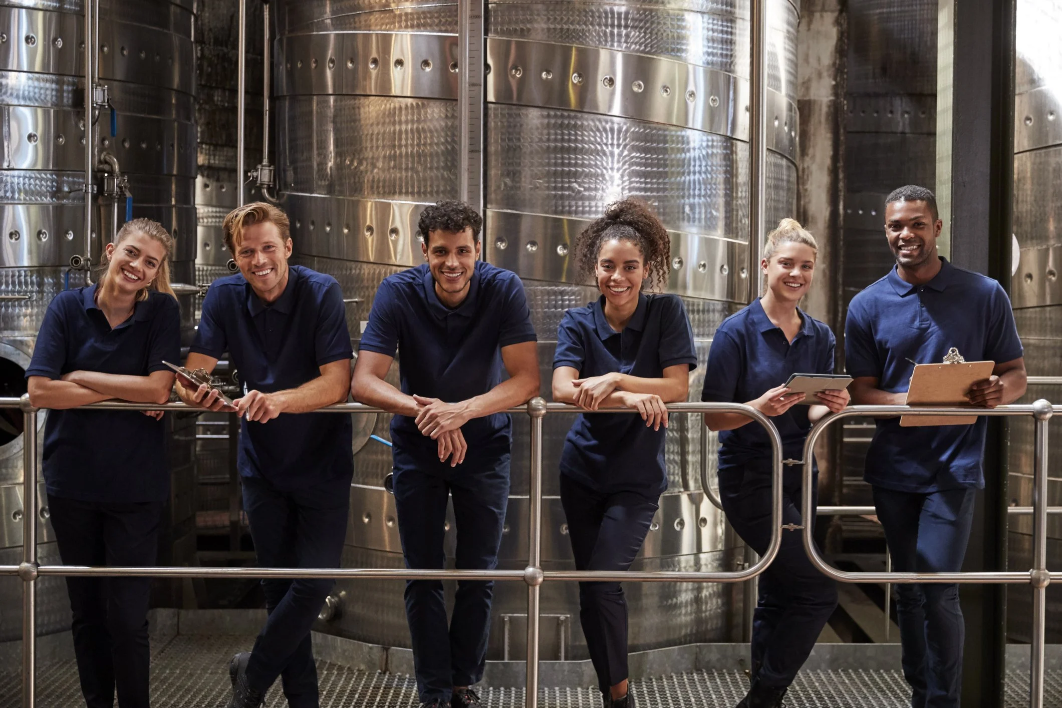 Six diverse workers in navy uniforms smiling and posing in a brewery or industrial setting with large metal tanks in the background.