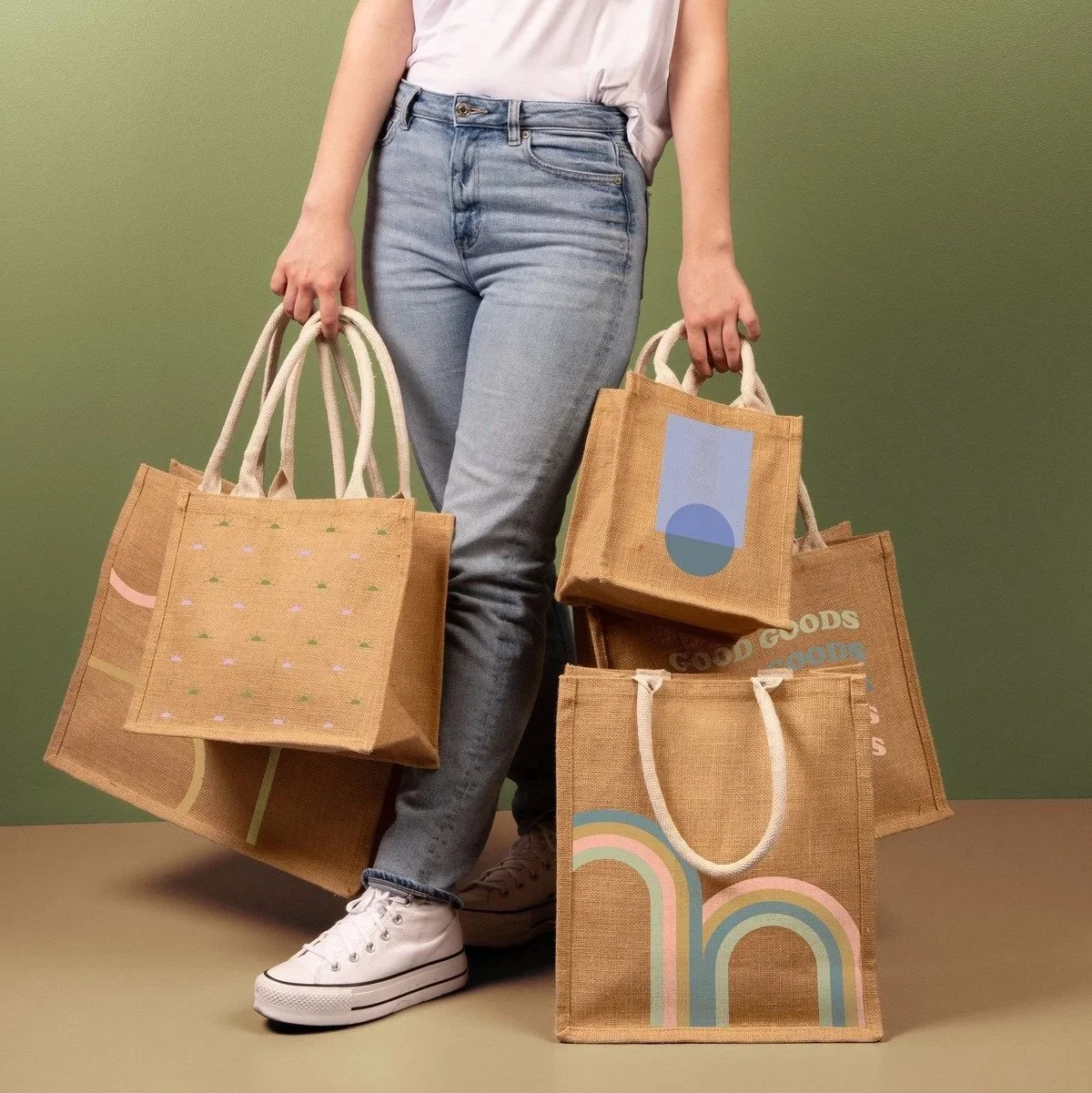 Person holding multiple reusable shopping bags, standing against a green background, wearing light blue jeans and white sneakers.