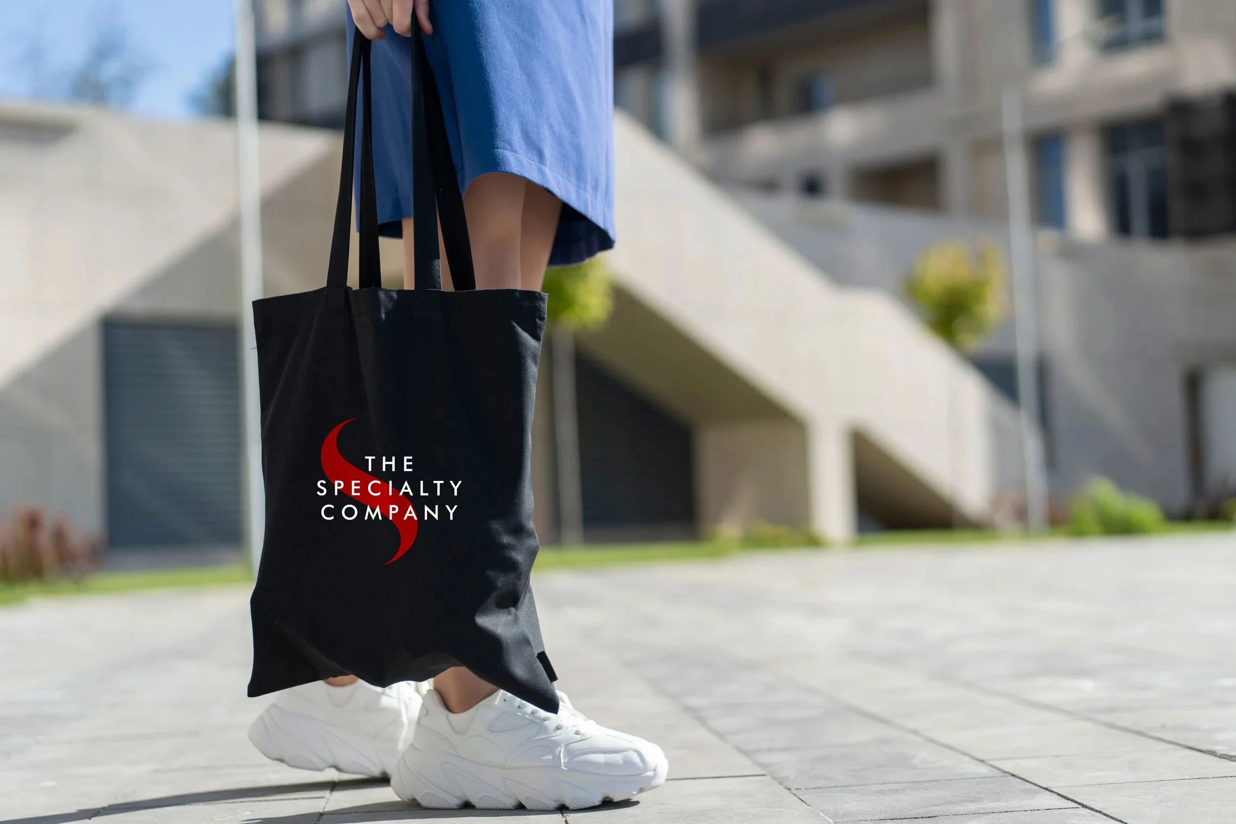 Person walking outdoors in a city area, wearing a blue dress, white sneakers, and carrying a black tote bag with red and white logo reading 'The Specialty Company,' with modern apartment buildings and trees in the background.