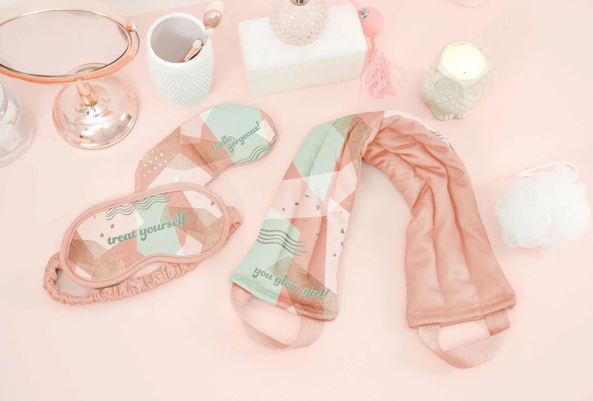 A pink-themed vanity or display table with a pink curtain, a pink eye mask reading 'treat yourself!', decorative candles, and a white and pink patterned scarf or cloth.