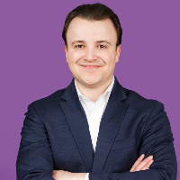 A man with short dark hair and light skin, wearing a navy blazer over a white shirt, standing with arms crossed against a purple background.