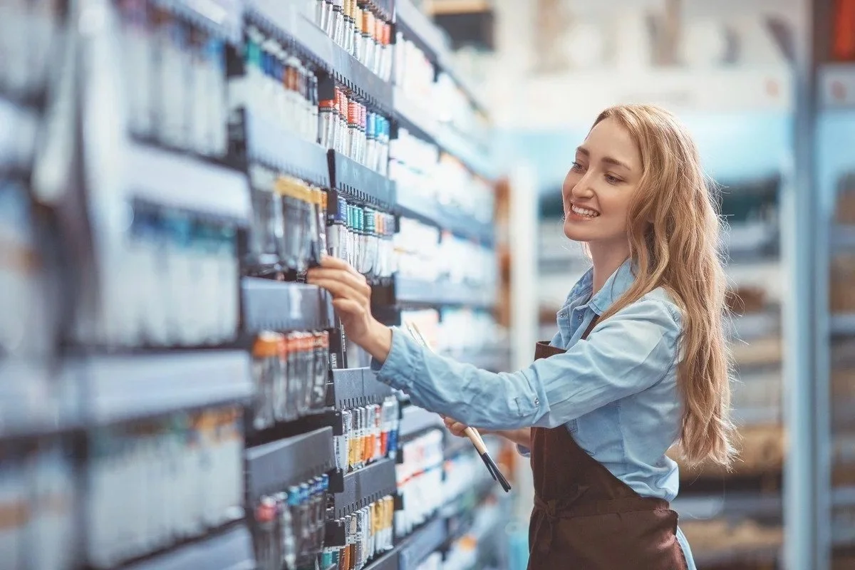 A young woman with long blonde hair shopping for paint in a hardware store, holding a clipboard and smiling as she picks a color from the shelves.