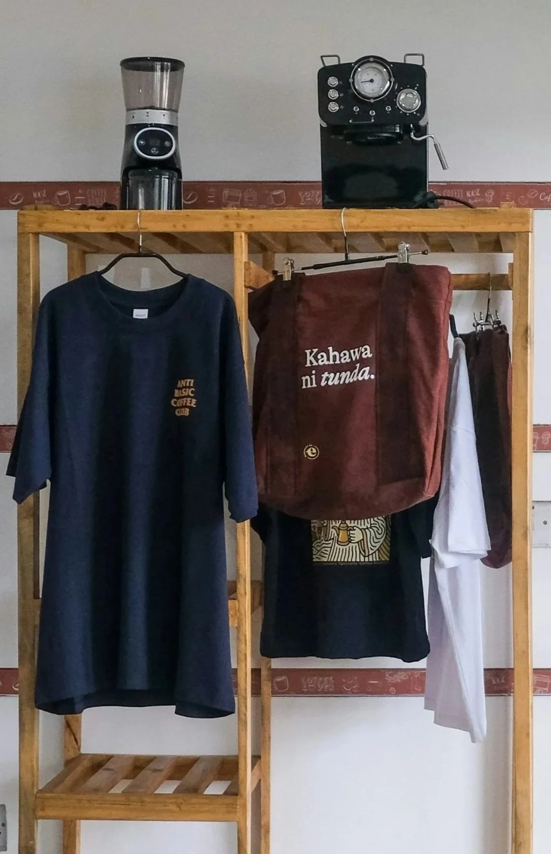 Wooden shelving unit with a coffee grinder and a vintage coffee machine on top, a navy blue T-shirt hanging on the left, a maroon tote bag with white text, and a white shirt hanging on the right.