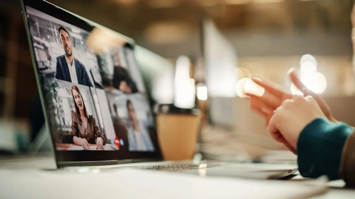 Person gesturing in front of a laptop screen showing a video call with multiple participants.