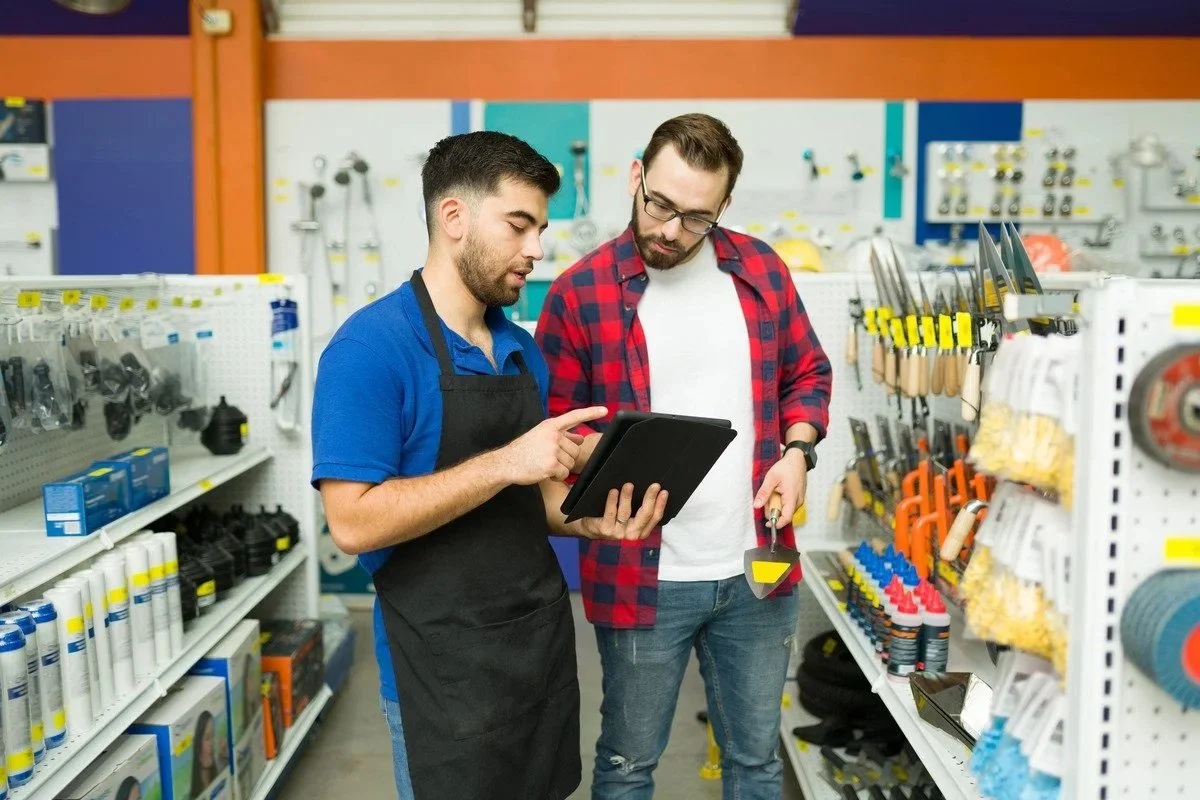 Two men in hardware store shop discussing tools, one worker with black apron and another customer with plaid shirt holding a screwdriver, looking at a tablet.