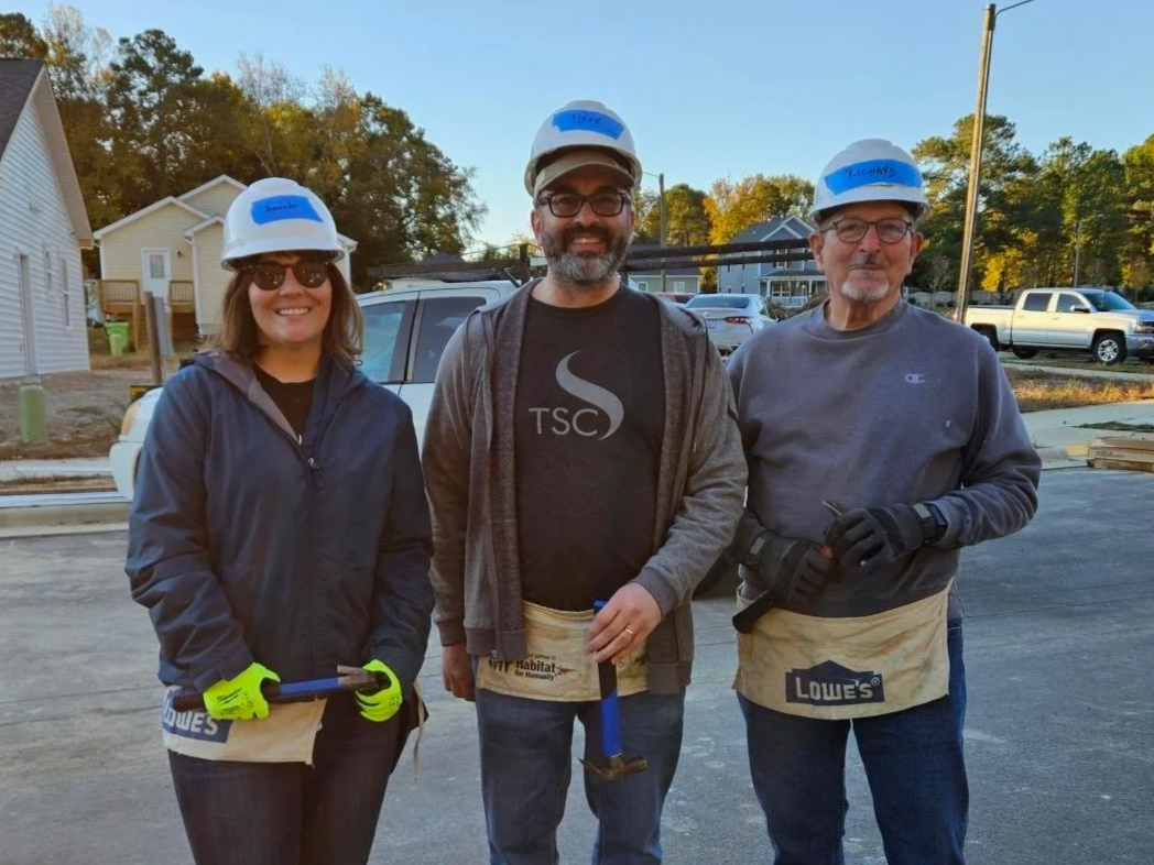 Three people wearing hard hats and gloves standing on a construction site during daylight, smiling at the camera.