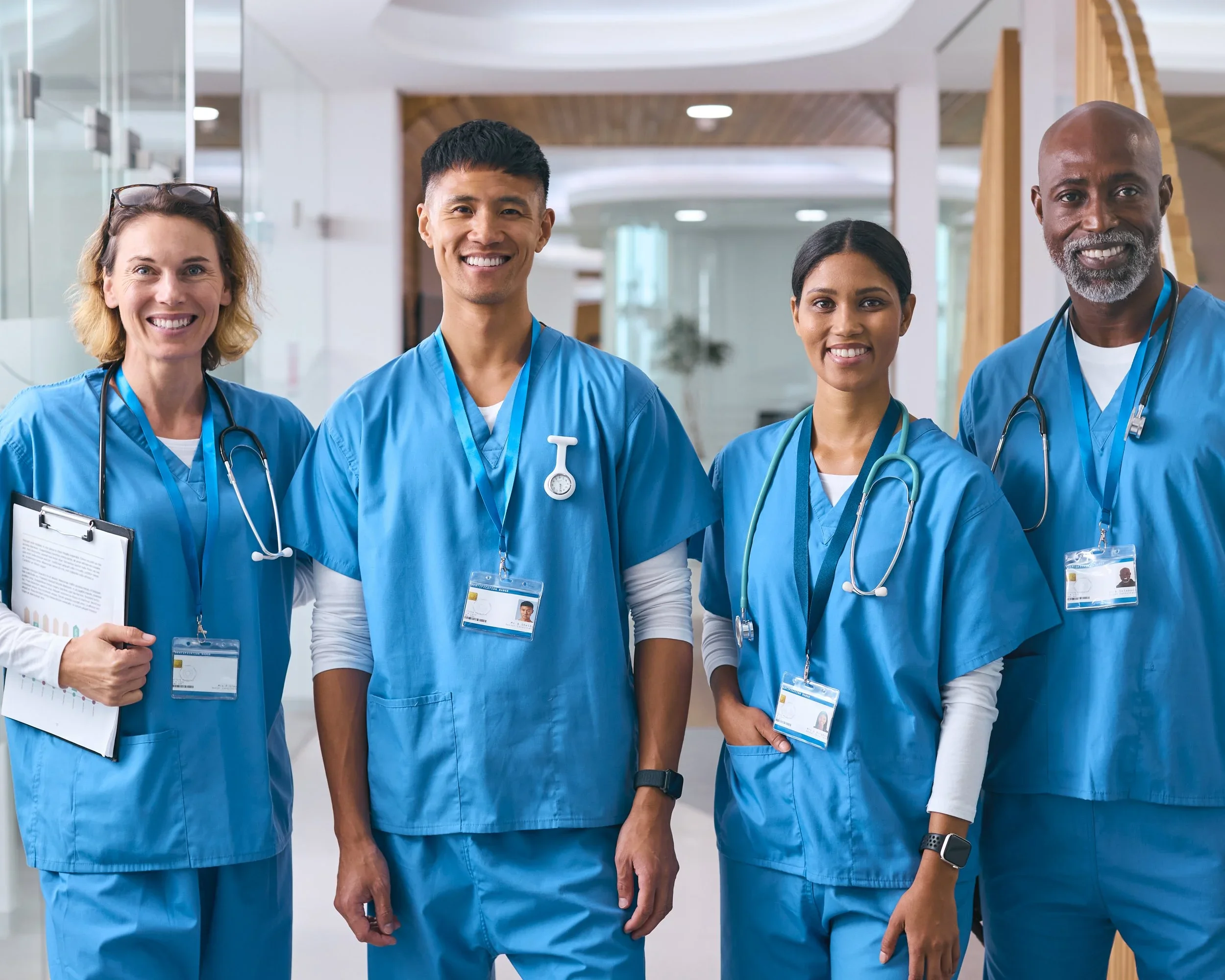 Four healthcare professionals in blue scrubs and stethoscopes smiling and standing in a hospital hallway.