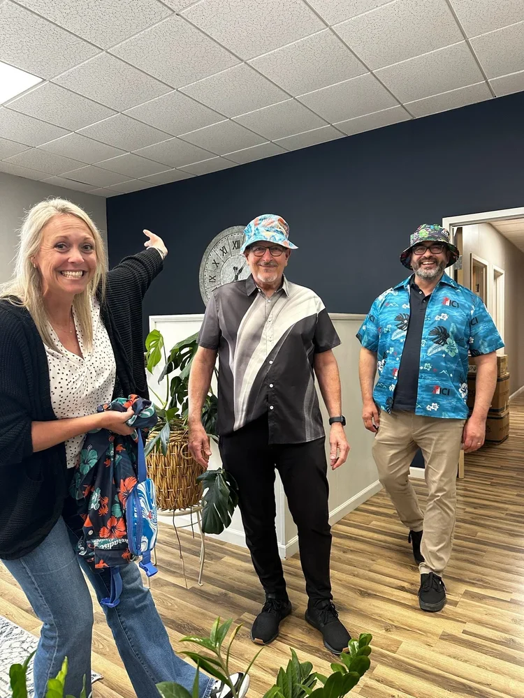 Three people standing indoors near a wall clock, with plants in the foreground. The woman on the left is smiling and wearing a black cardigan, white polka dot blouse, and jeans, holding a backpack. The man in the middle has glasses, a multi-colored bucket hat, and a short-sleeved shirt with black and gray diagonal color blocks. The man on the right is wearing a colorful floral Hawaiian shirt, tan pants, glasses, and a hat, smiling.