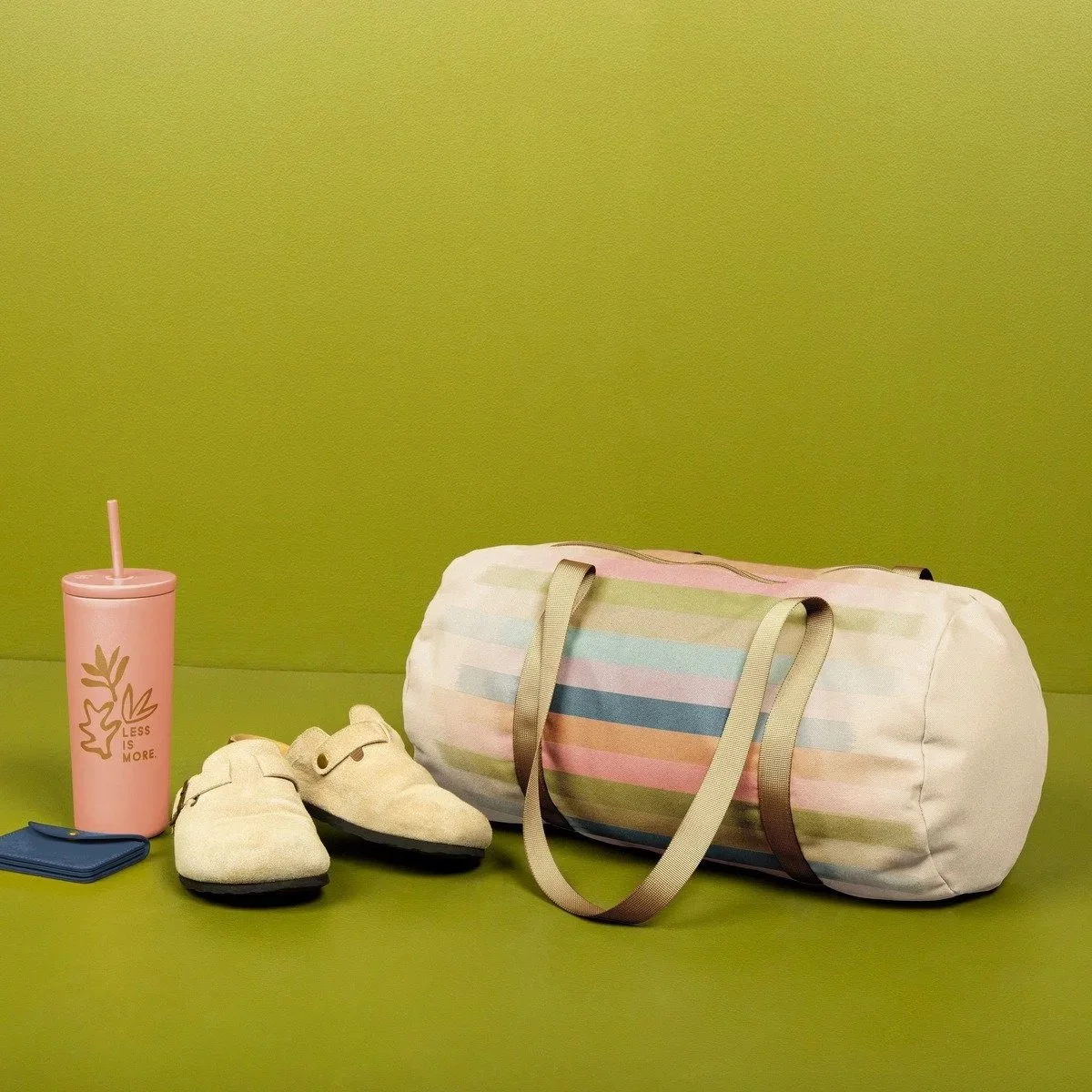 A collection of travel items on a green surface with a green background, including a pink reusable bottle with a straw, a pair of beige slippers, a small blue wallet, and a striped duffle bag with pastel colors.