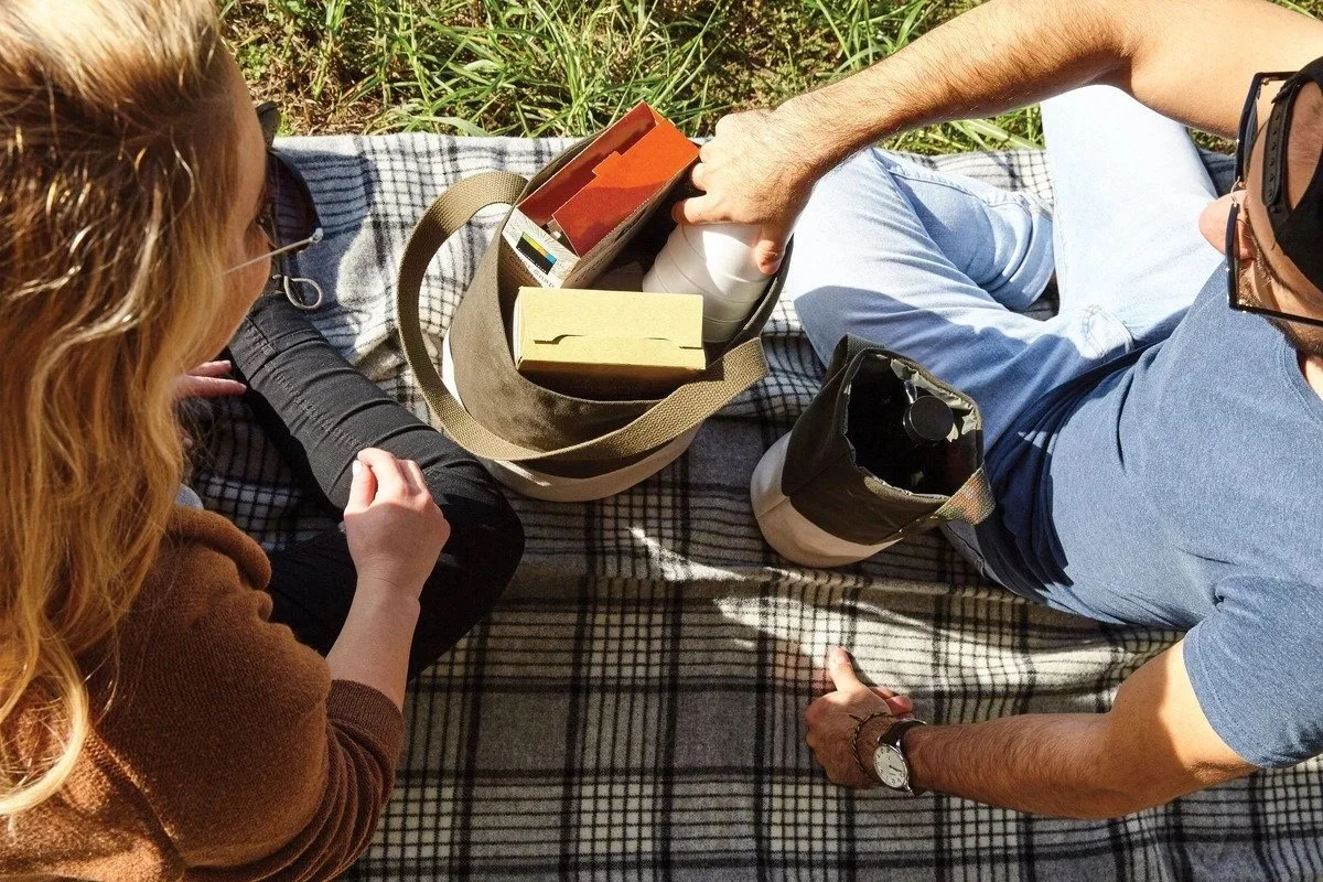 A man and woman having a picnic outdoors on a checkered blanket. The man is reaching into a tote bag with various items, and the woman looks on, sitting close together.