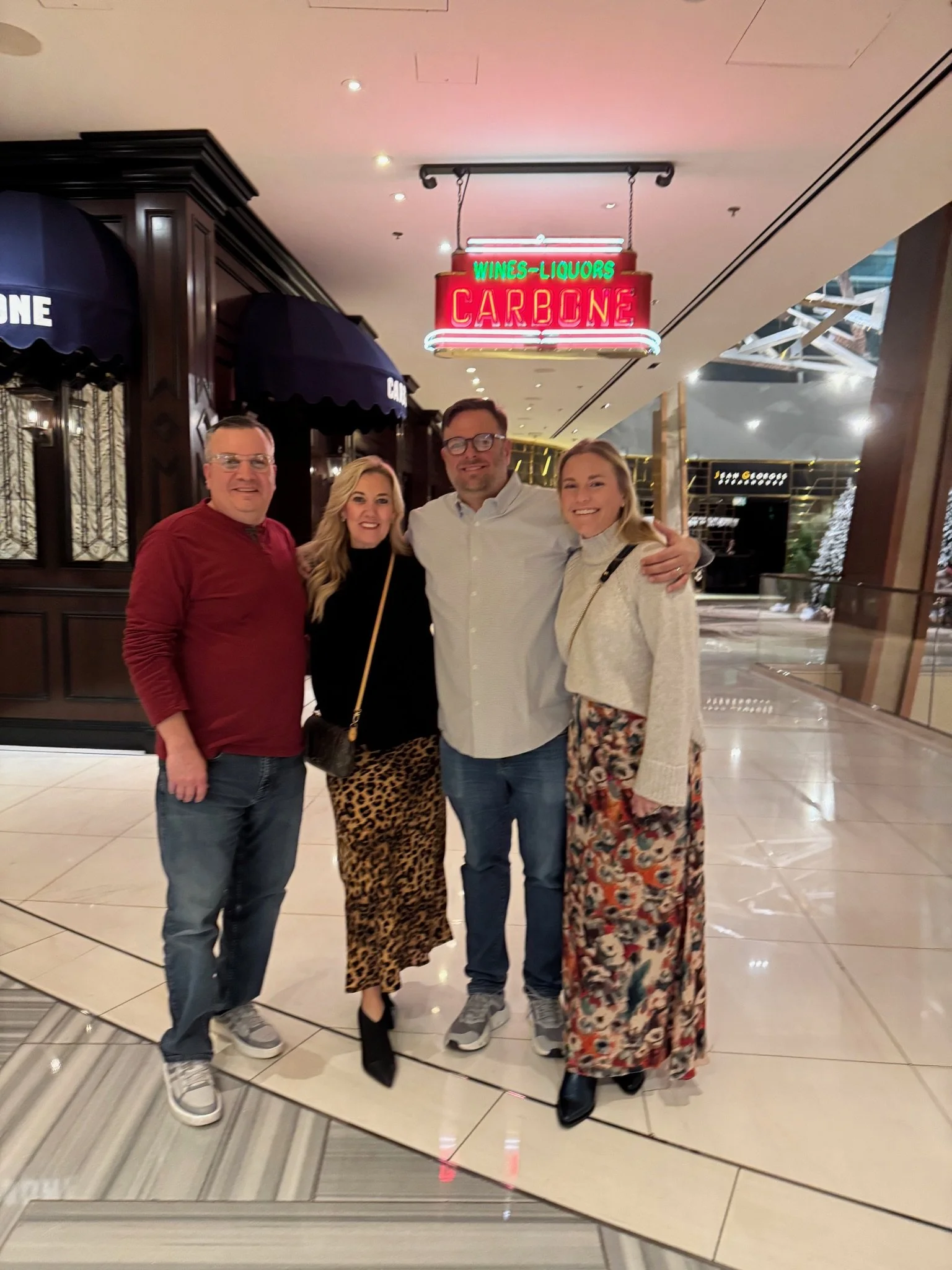 Four people posing in front of a neon sign that reads 'Wines - Liquors Carbone' inside a mall or hotel.