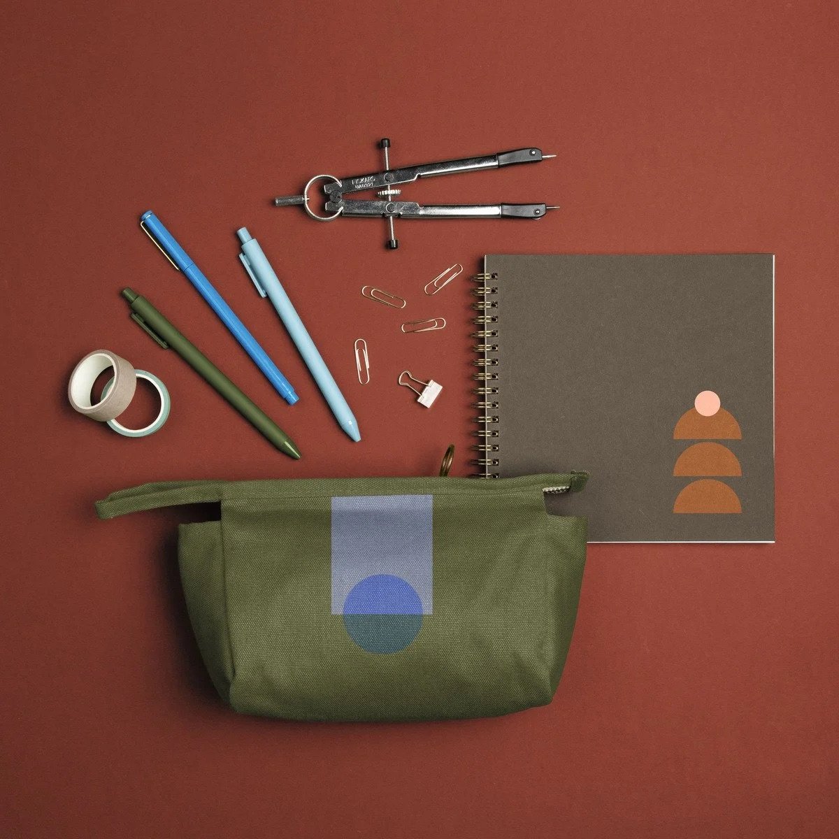 A flat lay of office supplies on a red background, including a green zippered pouch, a gray spiral notebook, a compass, pens, paper clips, washi tape, and a small white binder clip.