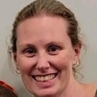 Close-up of a smiling woman with blonde hair pulled back, wearing a gray top, against a gray background.