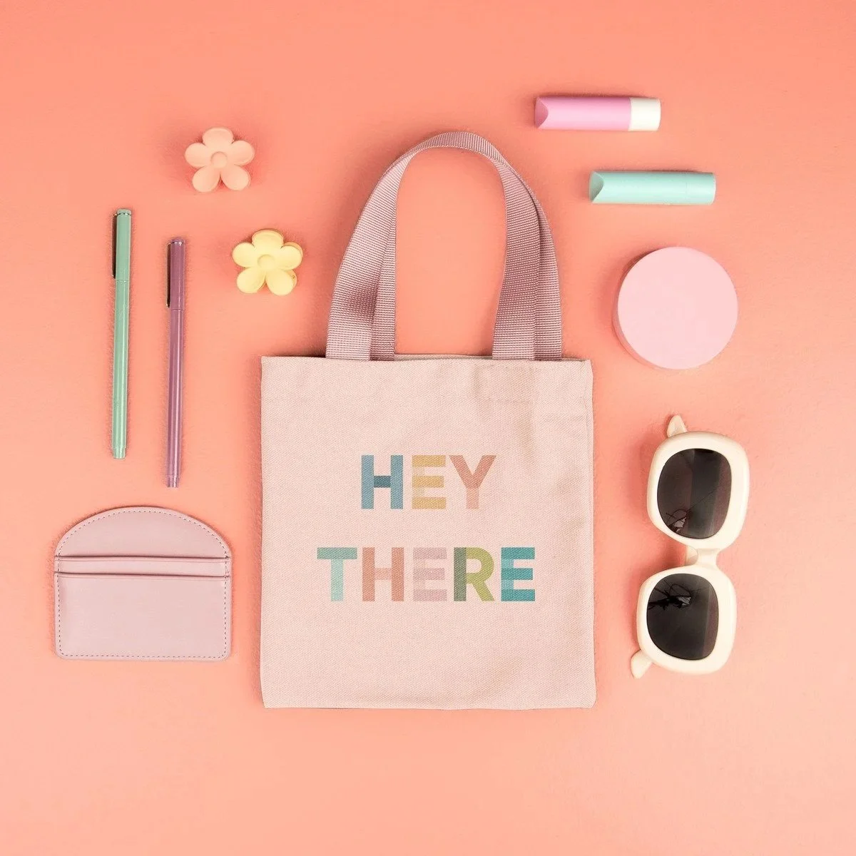 Flat lay of pastel-colored accessories and stationery around a tote bag with 'HEY THERE' written on it, set against a pink background. Items include sunglasses, highlighters, pens, flower-shaped erasers, a round container, and a small wallet.