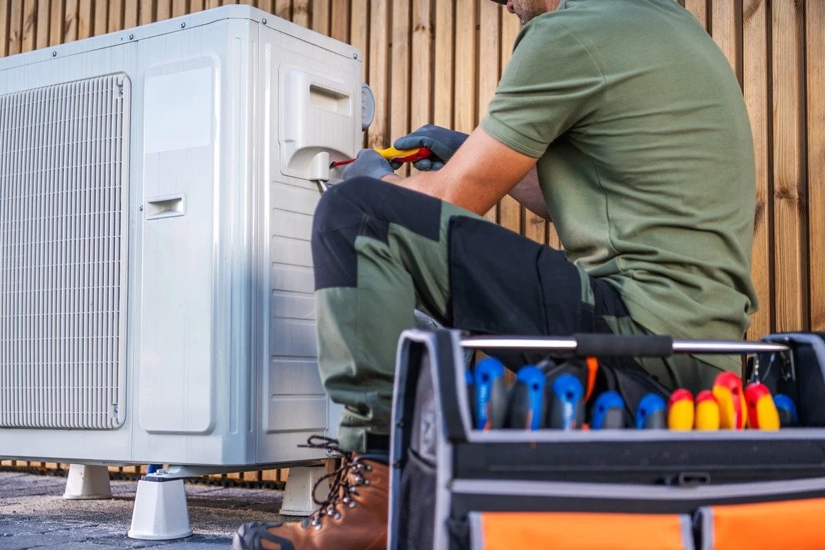 A technician in green work clothes and gloves repairing an outdoor air conditioning unit. There is a toolbox with screwdrivers nearby.
