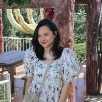A woman with dark hair wearing a white floral dress, smiling outdoors in a park with trees and wooden benches.