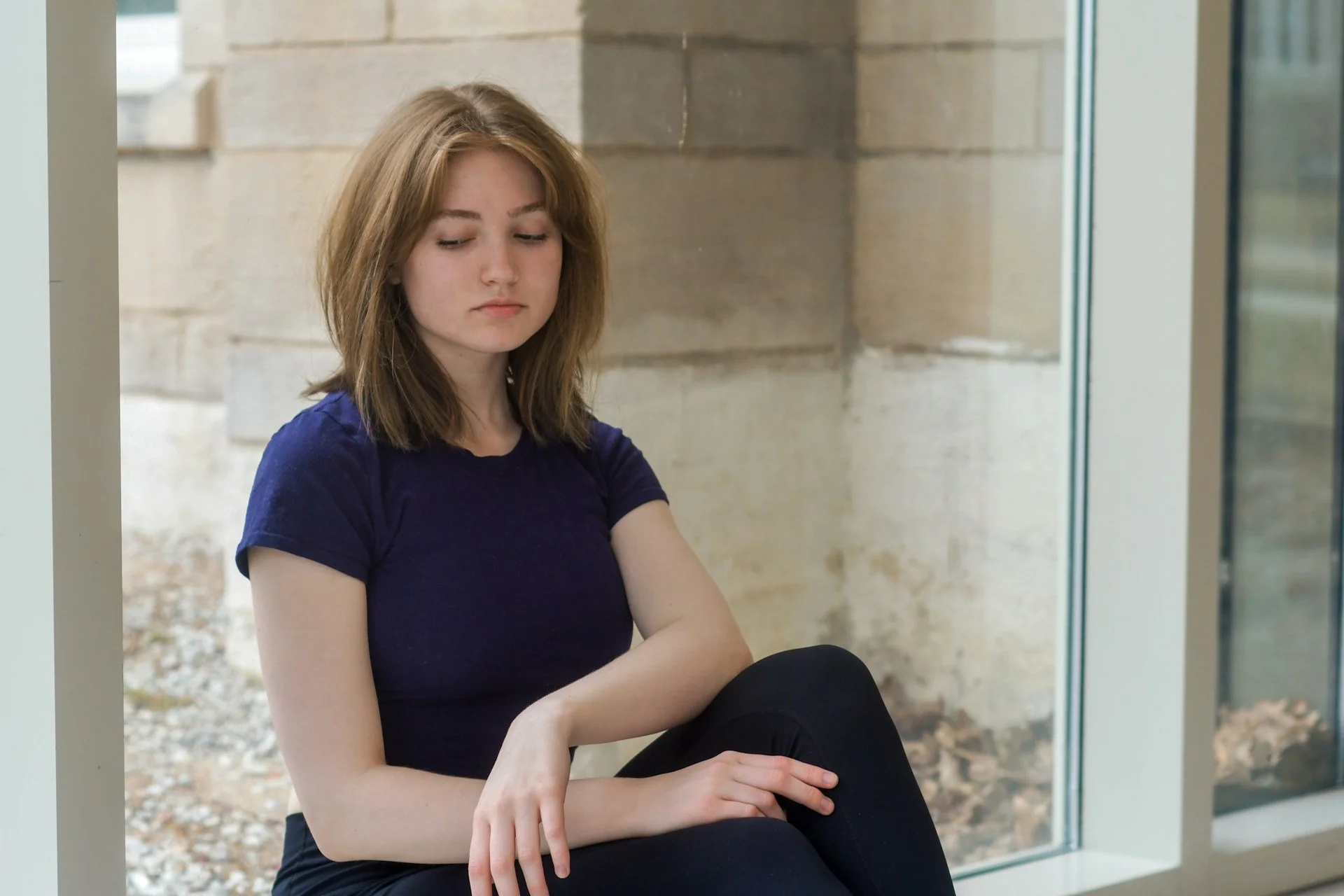 Young woman with brown hair wearing black pants and blue shirt sitting in front of a window
