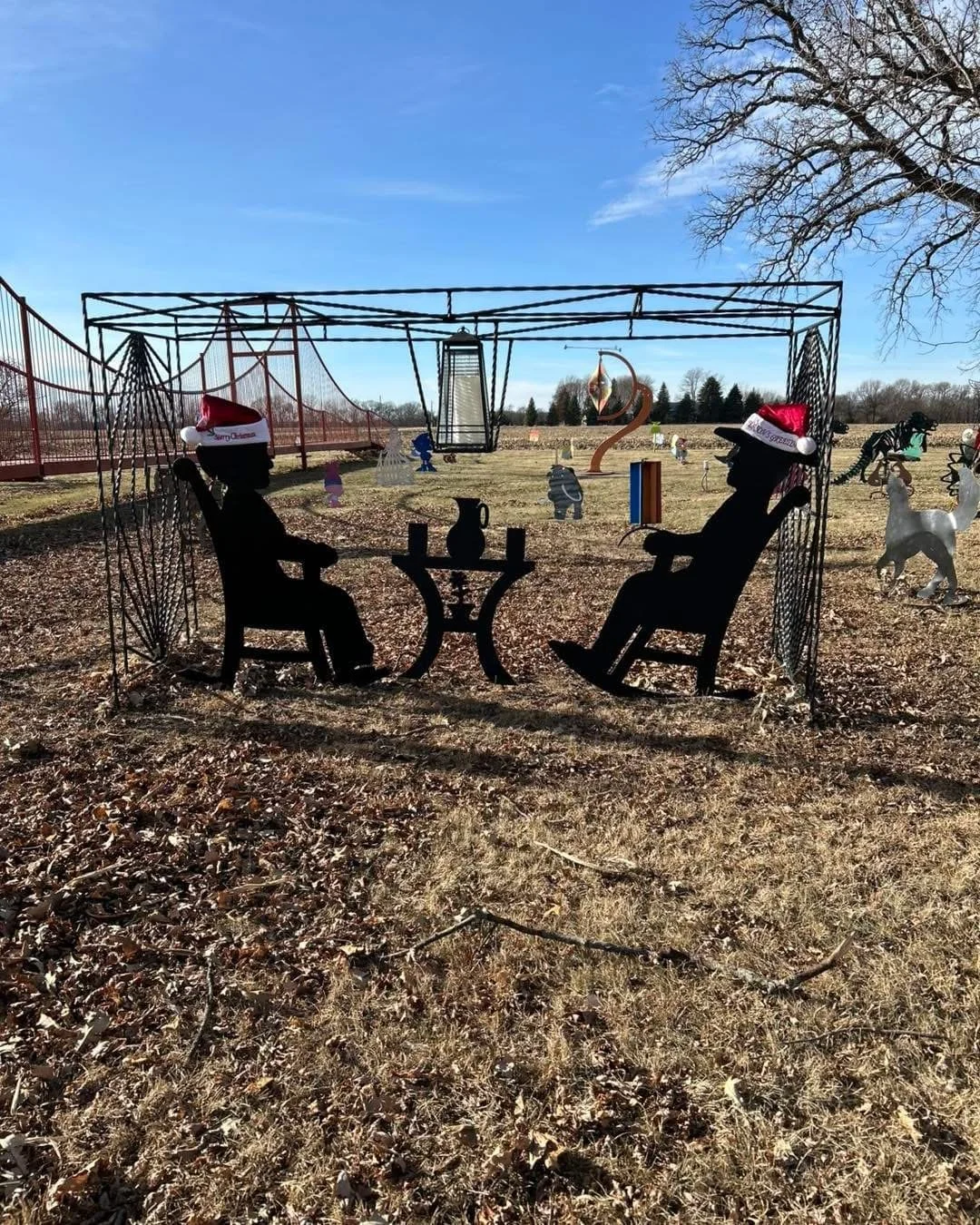 Silhouette of two people sitting at a table with drinks inside a wireframe gazebo, decorated with Santa hats, in a park, with sculptures, trees, and a bridge in the background on a bright, clear day.