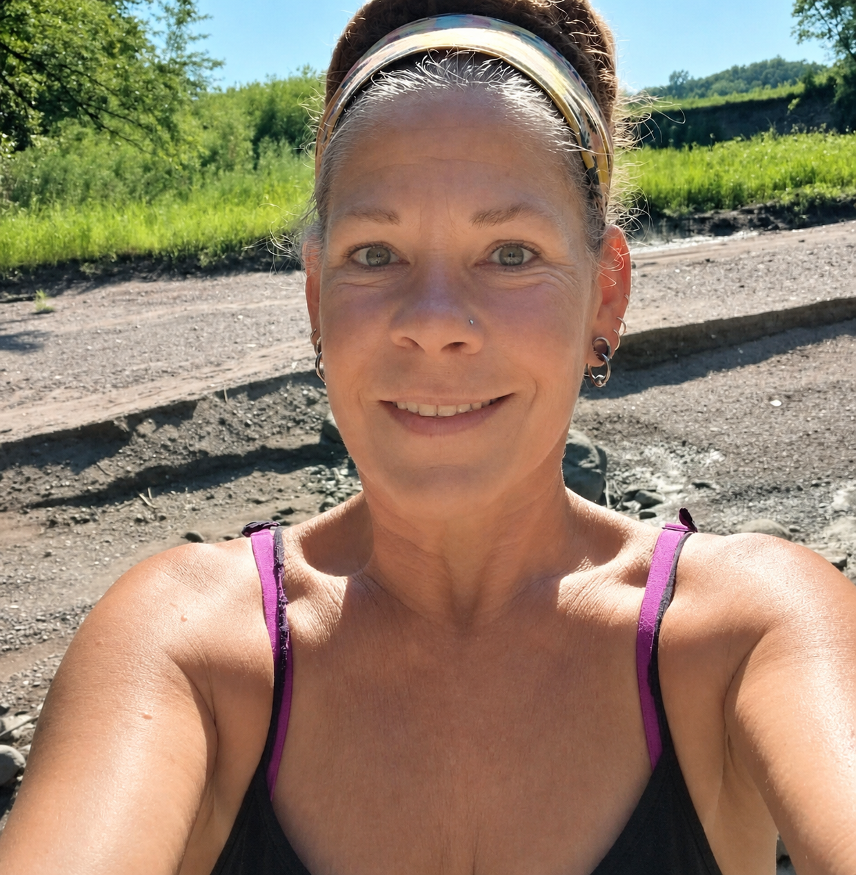 A woman smiling outdoors on a sunny day, wearing a headband, earrings, and a black top with purple straps, with trees and a dirt path in the background.