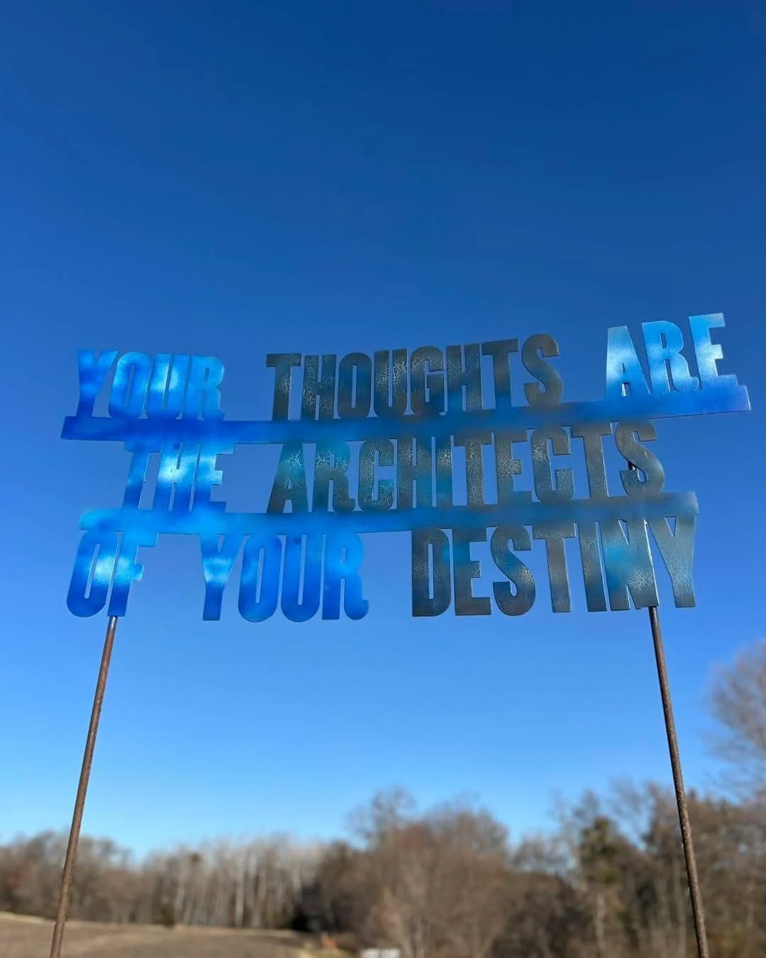 Metal sign with cut-out text that reads 'Your thoughts are the architects of your destiny,' held up by two metal rods against a clear blue sky with trees in the background.
