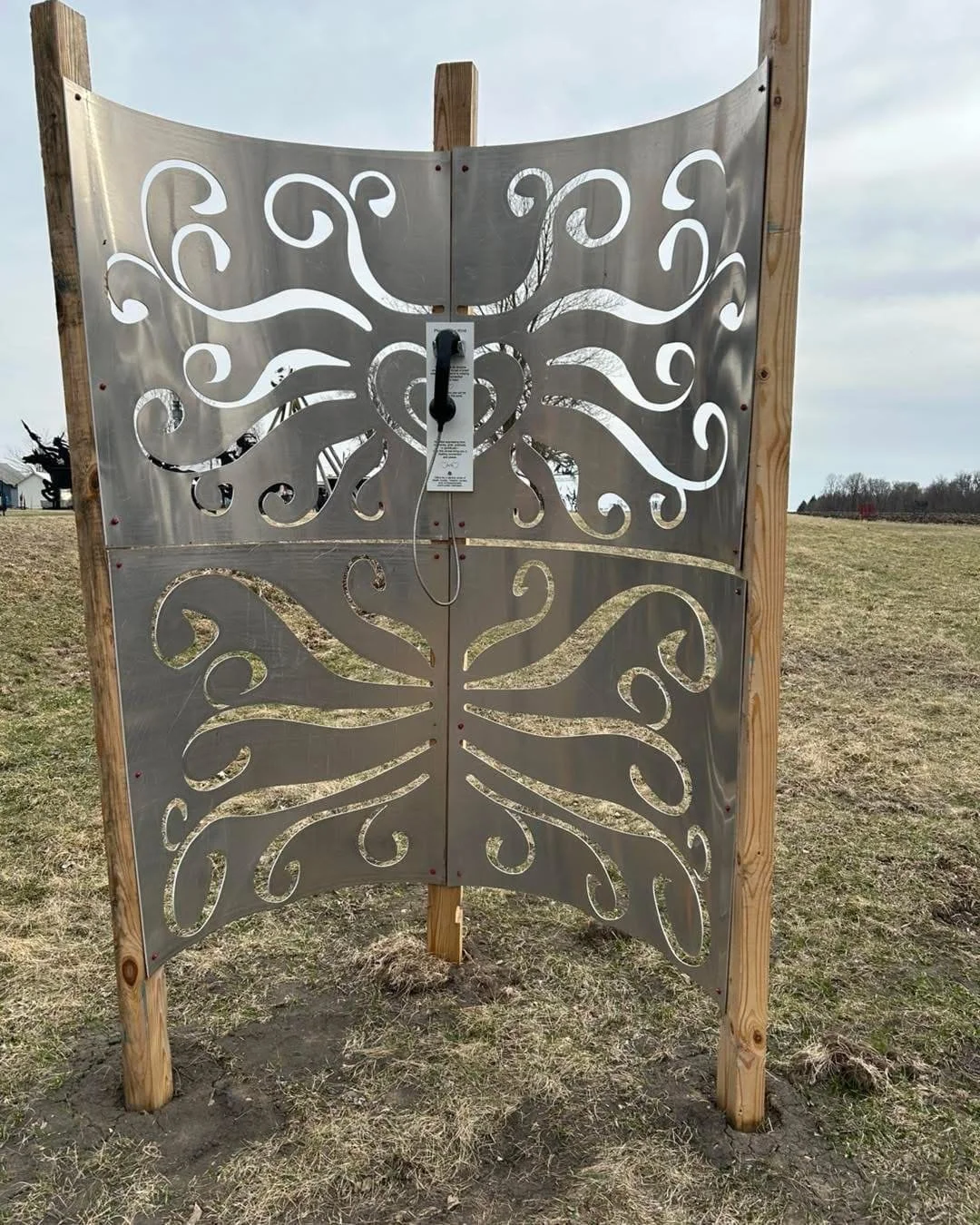 Outdoor installation with two metal panels featuring butterfly and floral cutout designs, mounted on wooden posts, in a grassy area.