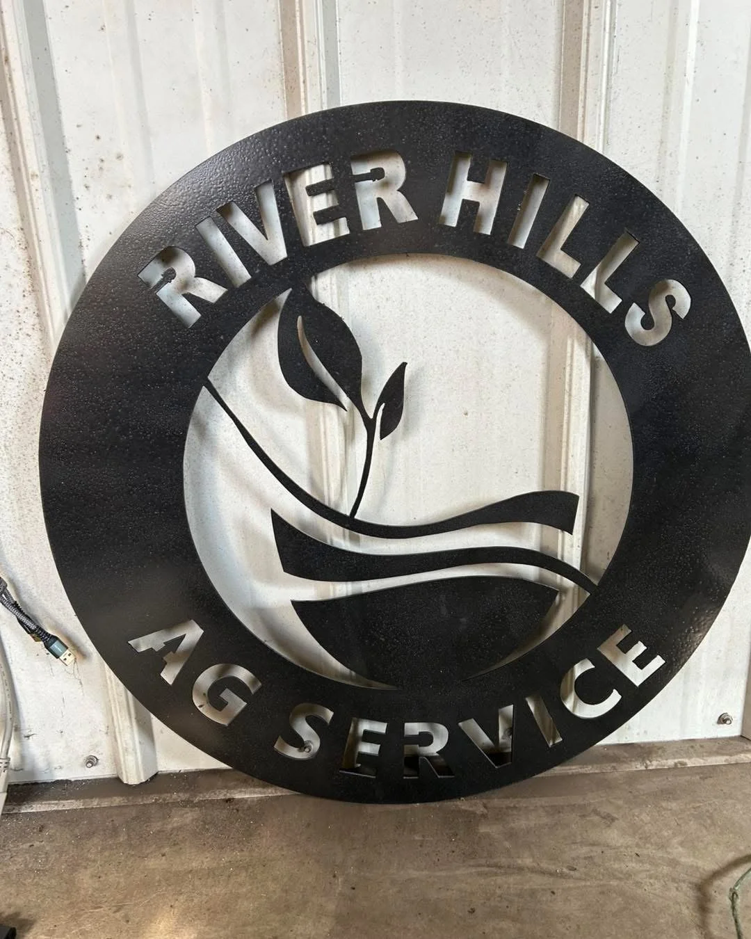 Black metal sign for River Hills Ag Service with a circular design and a plant symbol in the center, mounted on a white wall.