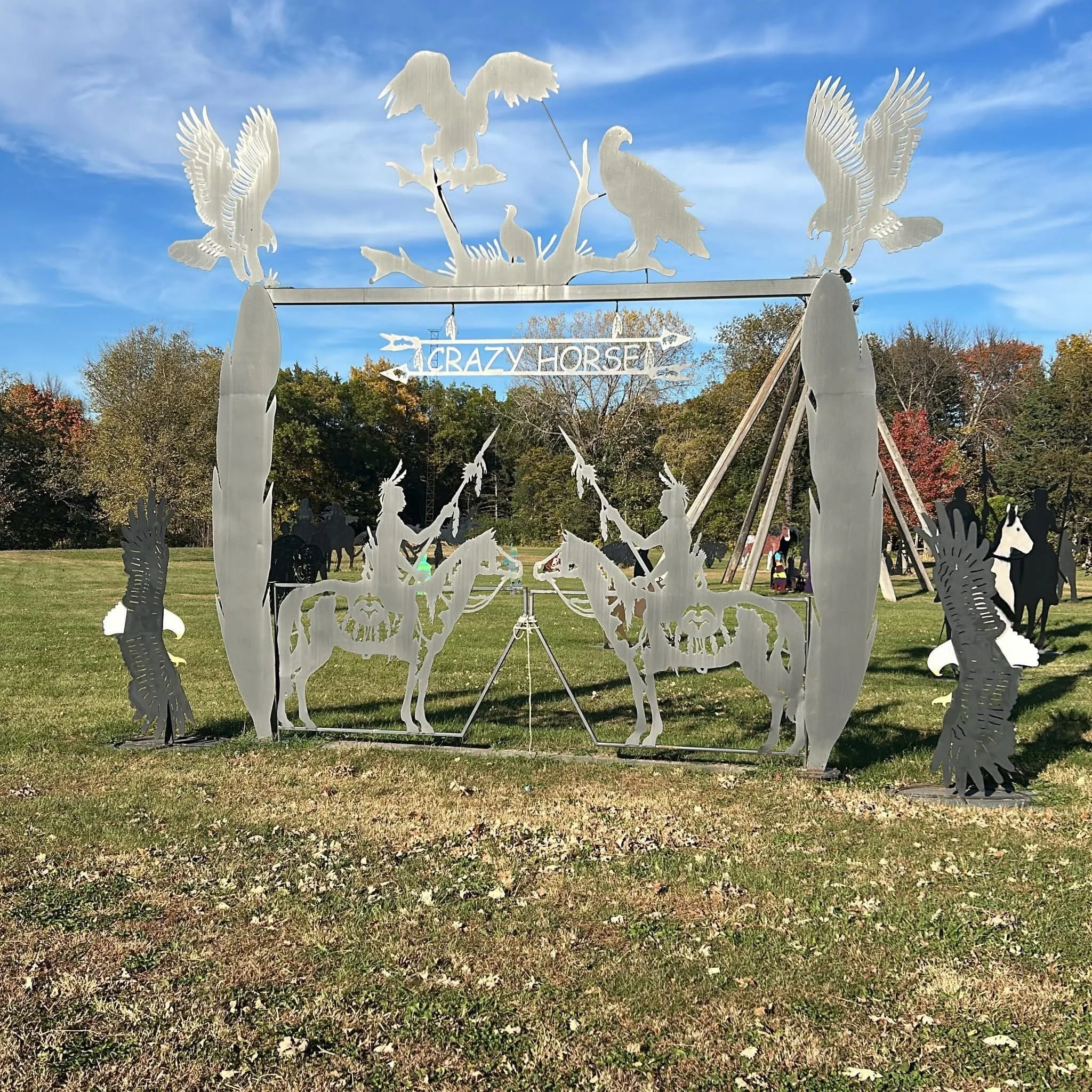 Metal sculpture of multiple birds, including eagles and a hawk, flying and perched, with the words 'Crazy Horse' on a sign below, set against a backdrop of trees and a blue sky.