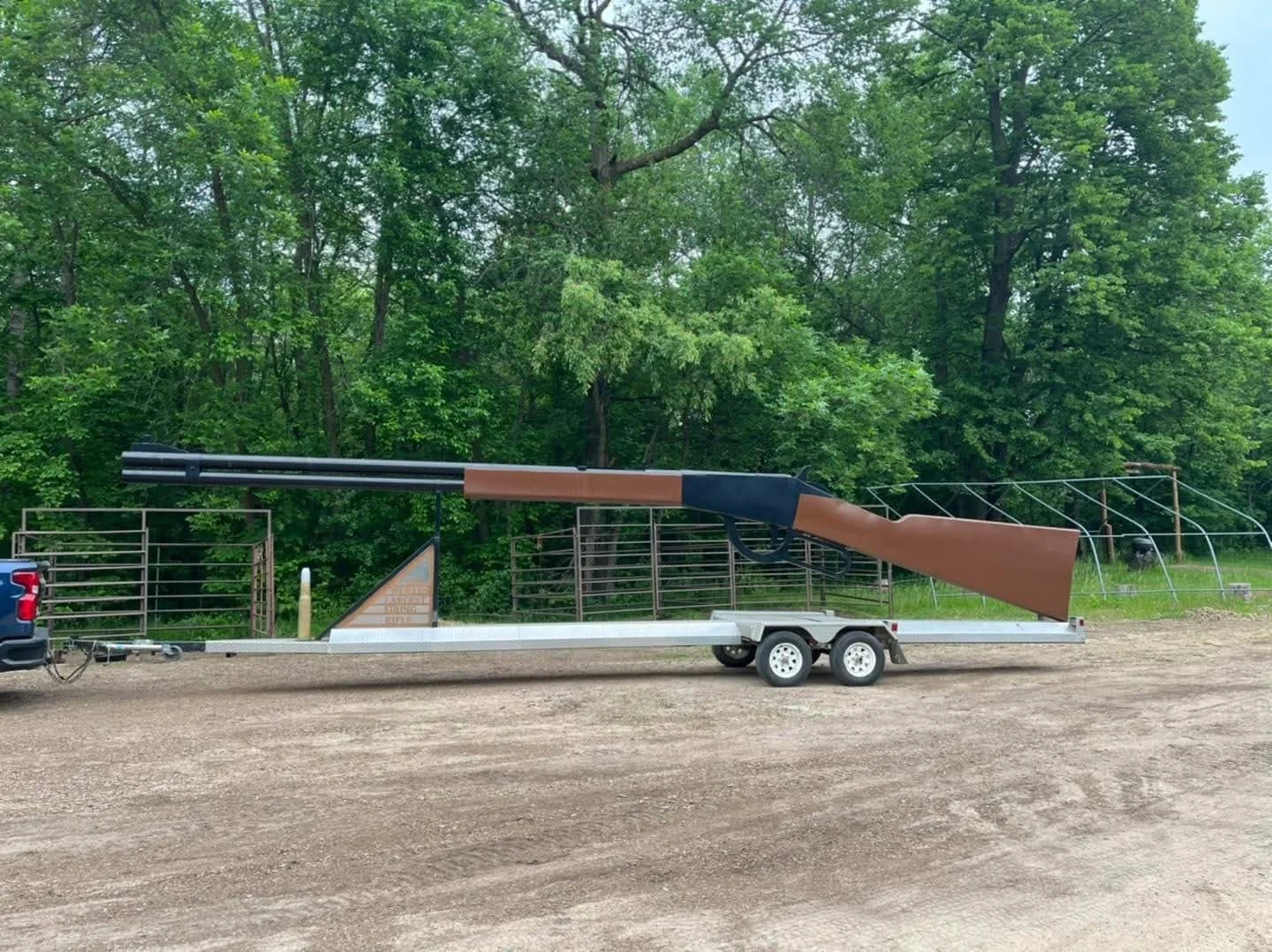 A large artificial rifle sculpture mounted on a trailer in front of a green wooded area.