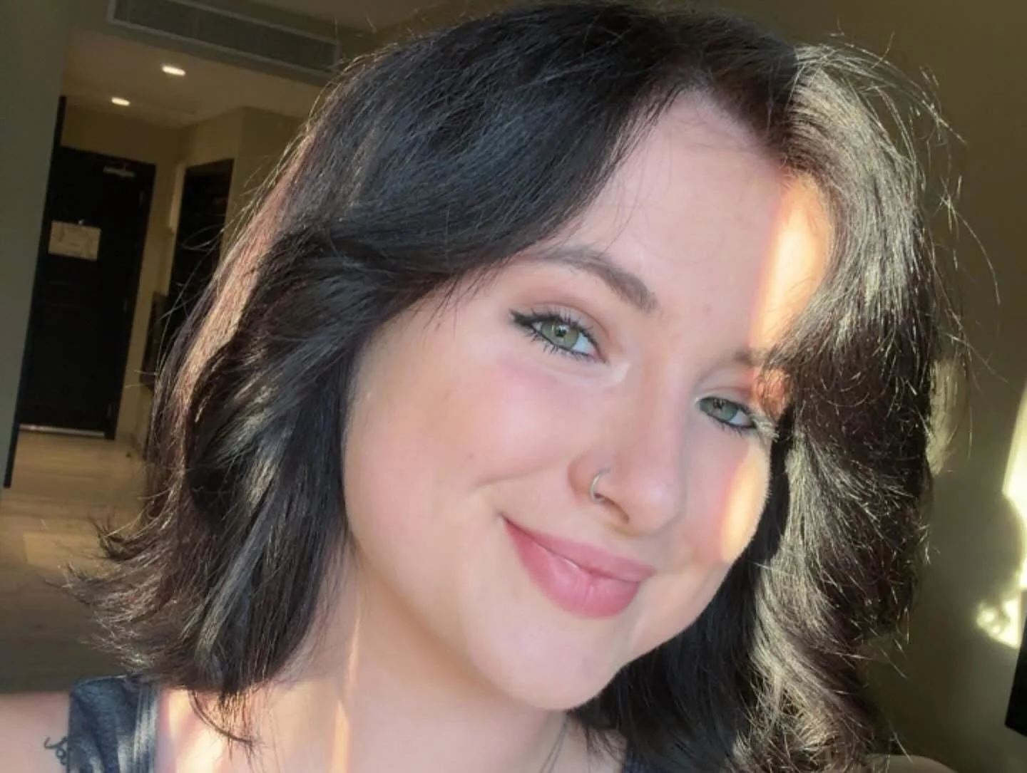 Close-up of a young woman with dark hair and green eyes smiling, with sunlight illuminating her face in an indoor setting.
