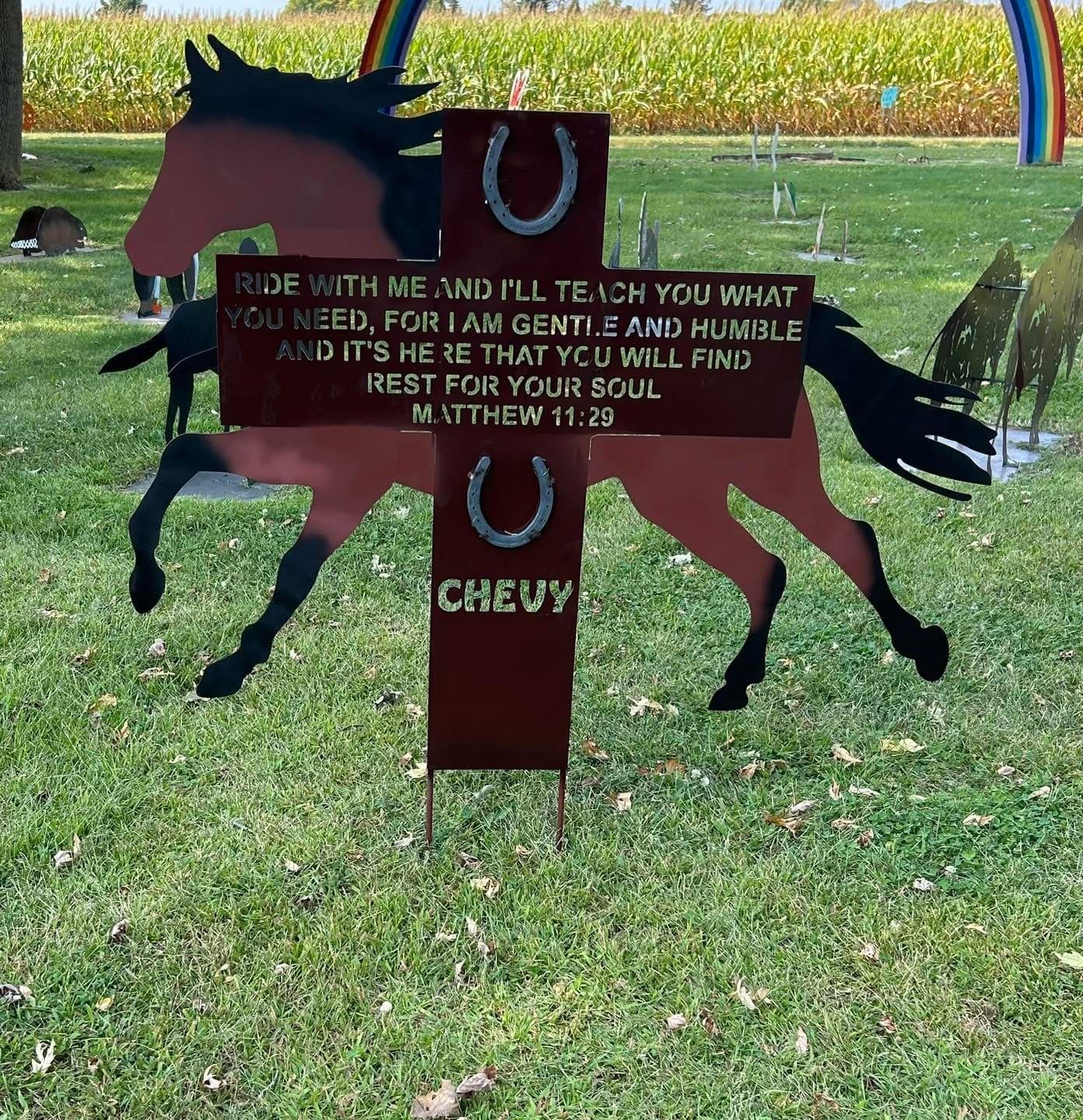 A decorative metal sign featuring a silhouette of a running horse, with a motivational quote about riding, Jesus, and rest for the soul. The sign includes horseshoes, a rainbow arch, and the word 'CHEWY' at the bottom, set on green grass with a farm 