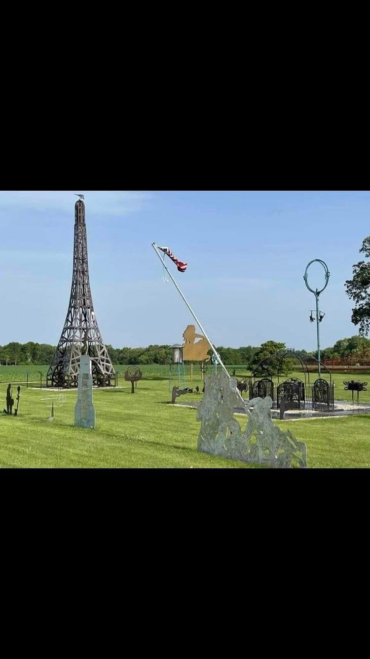 Outdoor scene with various sculpture art pieces on green grass, including a tall Eiffel Tower replica, a flagpole with American flag, and other abstract metallic sculptures, trees in background.