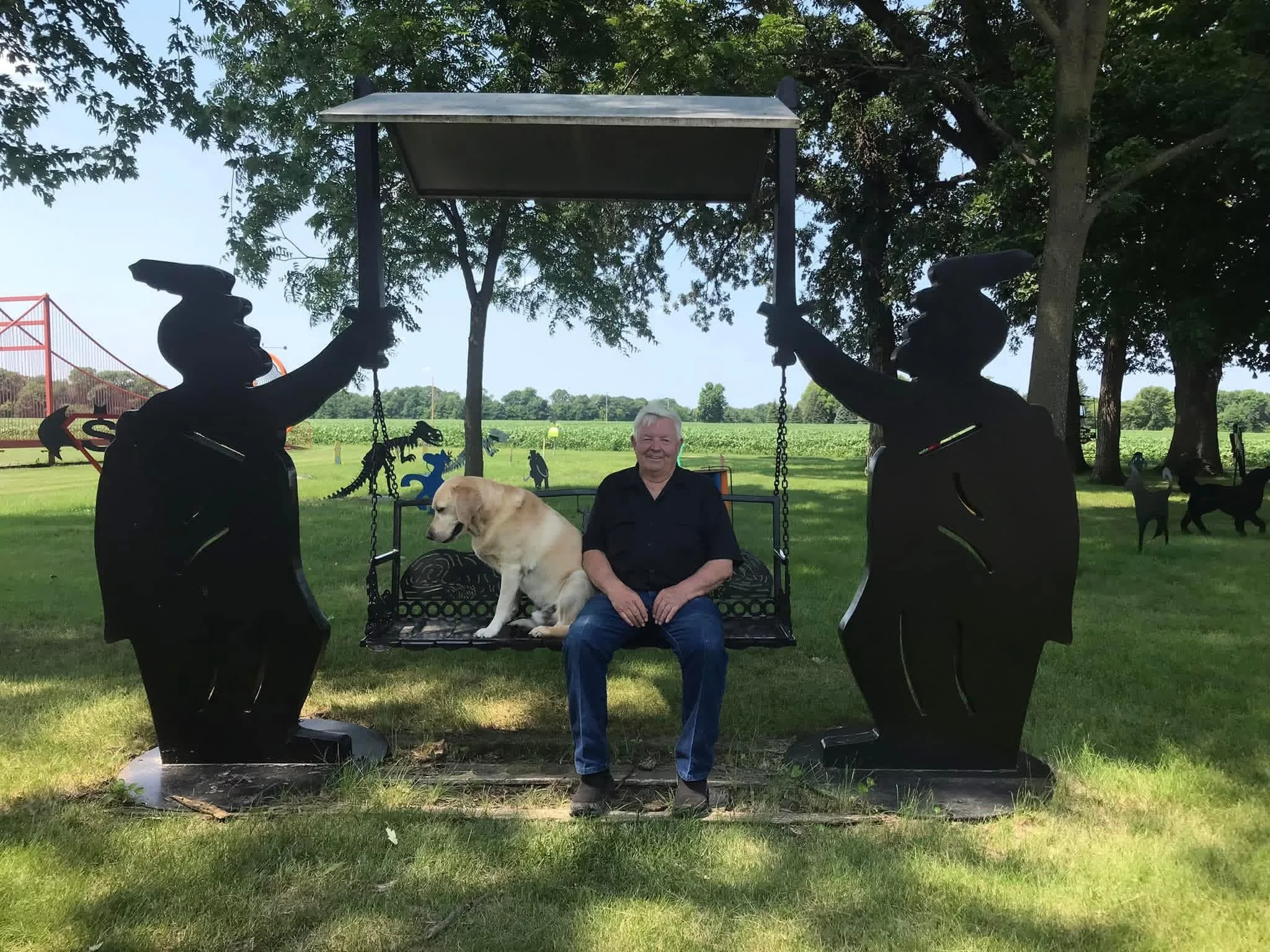 A person sitting on a bench with a yellow Labrador Retriever dog, surrounded by black dinosaur sculptures in a park with trees and open field in the background.