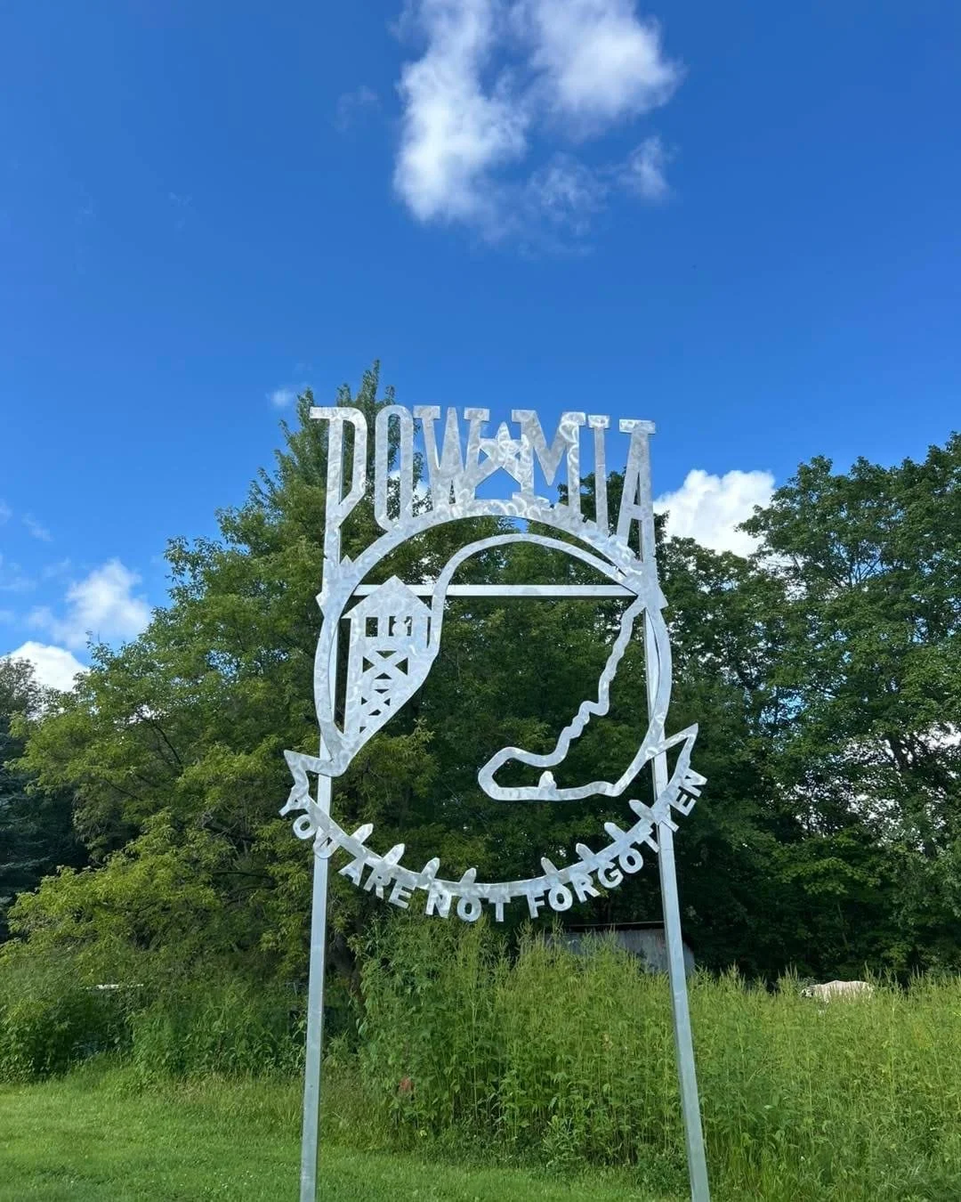 Metal sign with the text 'Plymouth' and a silhouette of a person's profile, surrounded by trees and a partly cloudy blue sky.