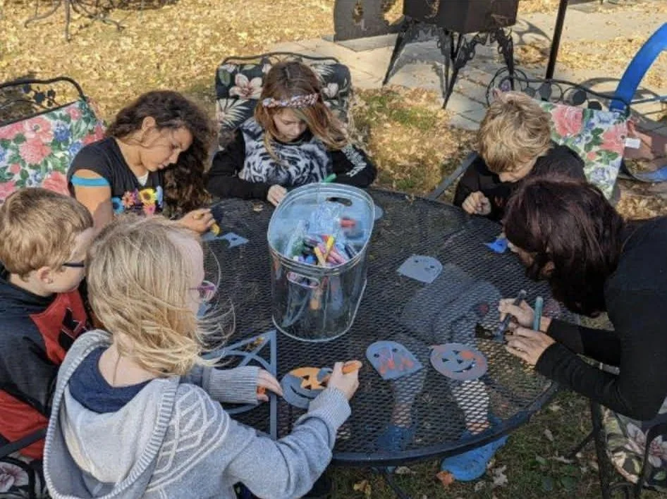 People, including children, sitting around a black metal outdoor table doing arts and crafts, making Halloween jack-o'-lantern decorations from paper.