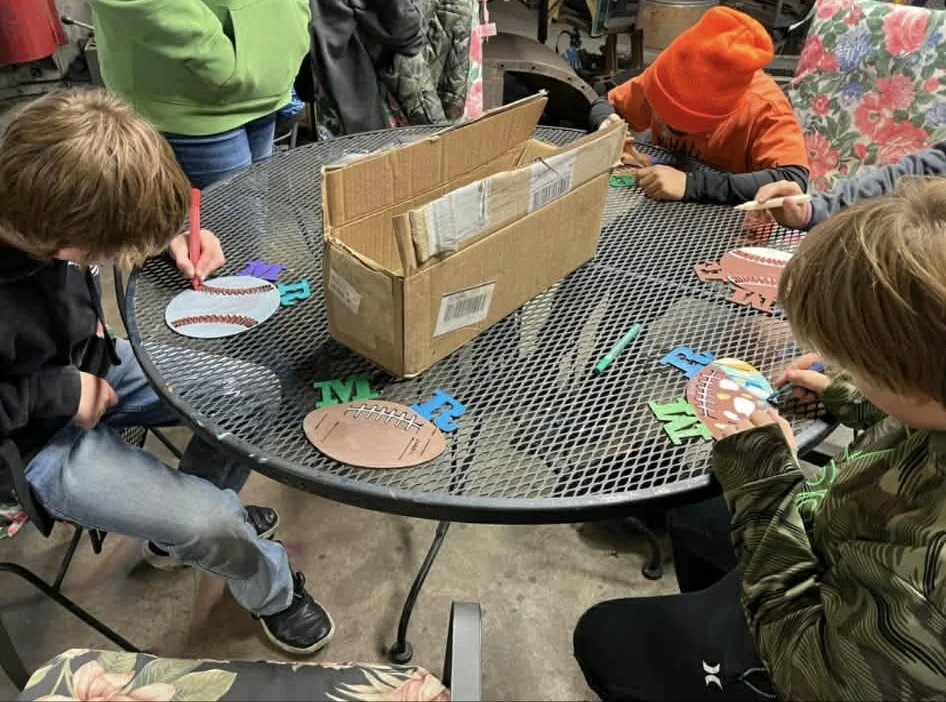 Children sitting around a round metal table, decorating football-themed paper cutouts with markers, in a workshop or craft area.