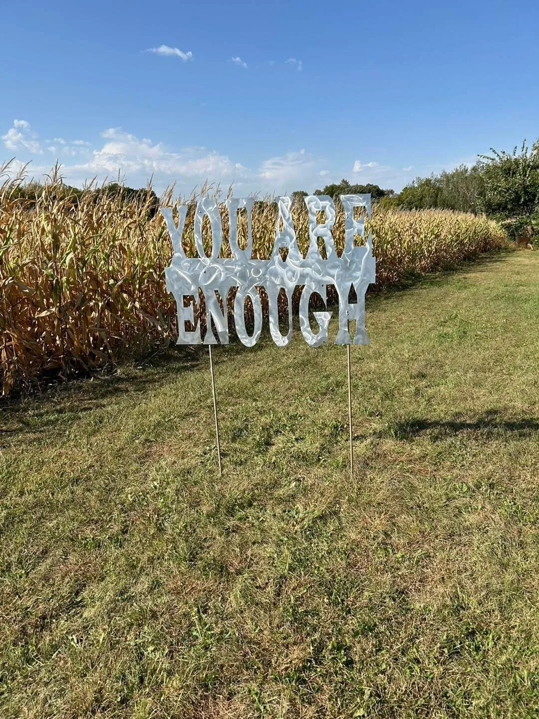 A white decorative sign with the words 'You're Enough' stands on two metal stakes in a grassy field, with a cornfield and a blue sky with some clouds in the background.