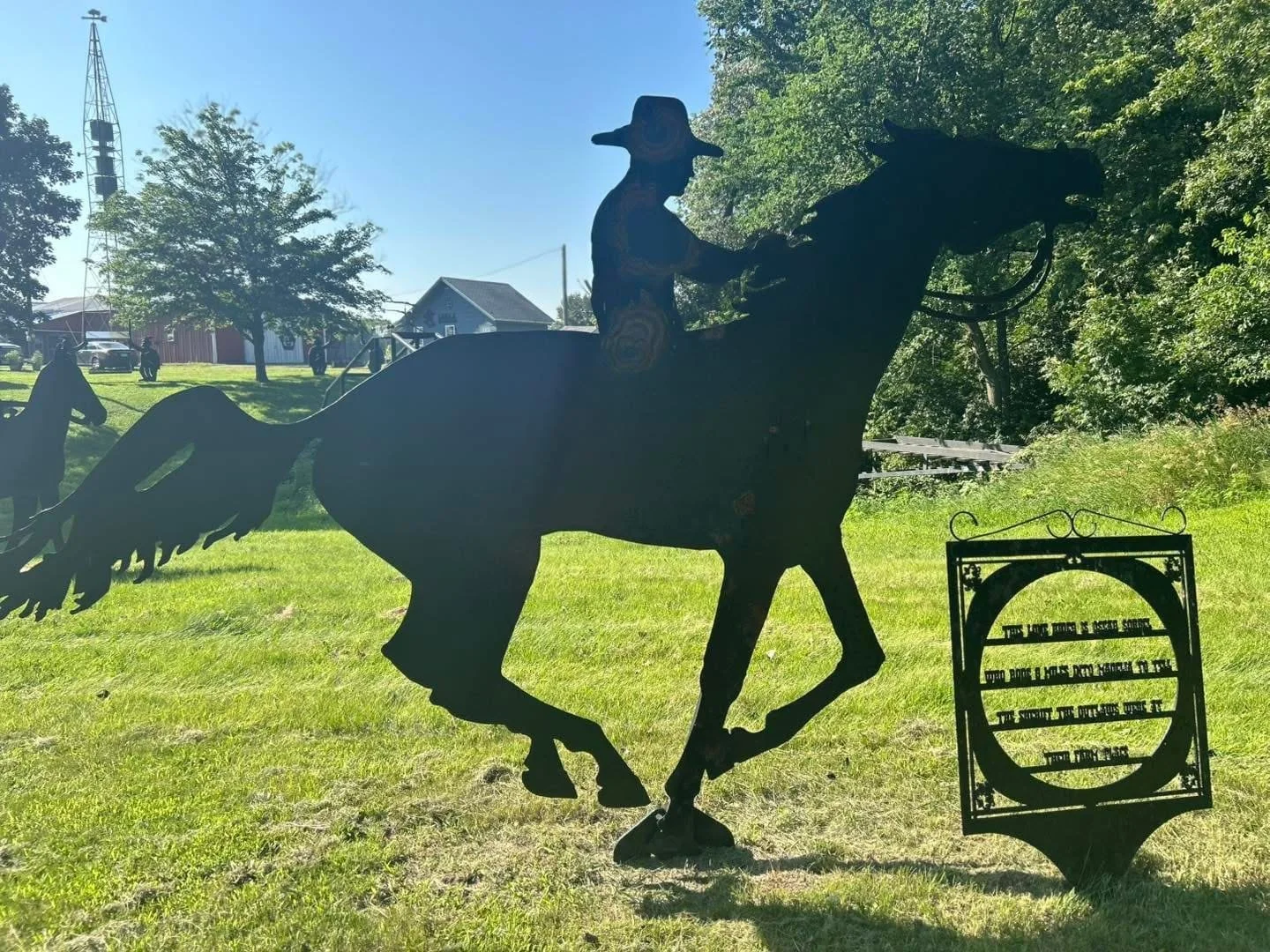 Metal silhouette sculpture of a cowboy riding a bucking horse, with trees and buildings in the background, situated outdoors on a grassy area.
