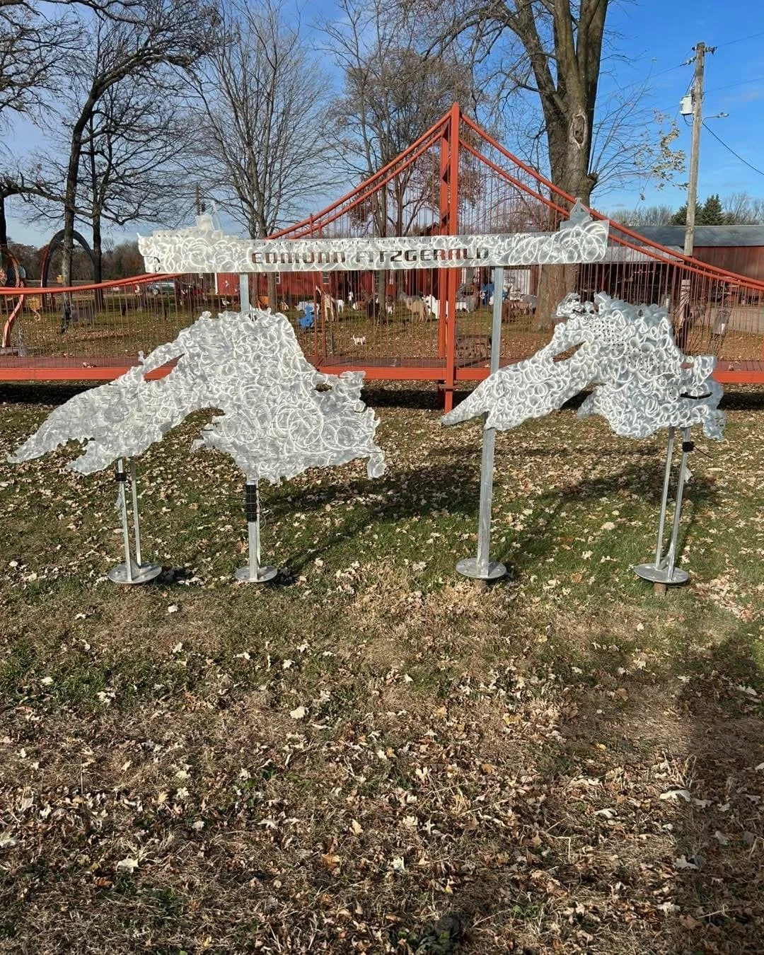 Decorative metal sculptures of white horses, mounted on bases, are displayed outdoors in front of a red suspension bridge with a sign that reads 'Edinburg ISD General' in an open area with trees and brown grass.