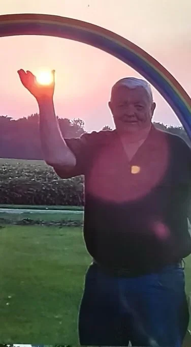 A person standing outdoors near a rainbow-shaped arch, holding the setting sun with their hand, with a background of trees and a field at sunset.