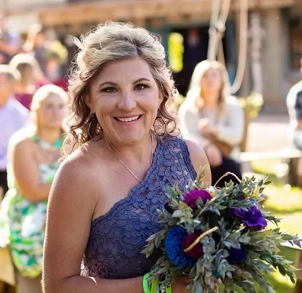 A woman with shoulder-length grayish-brown hair smiling and holding a bouquet of purple, green, and blue flowers at an outdoor event, with people and wooden structures in the background.