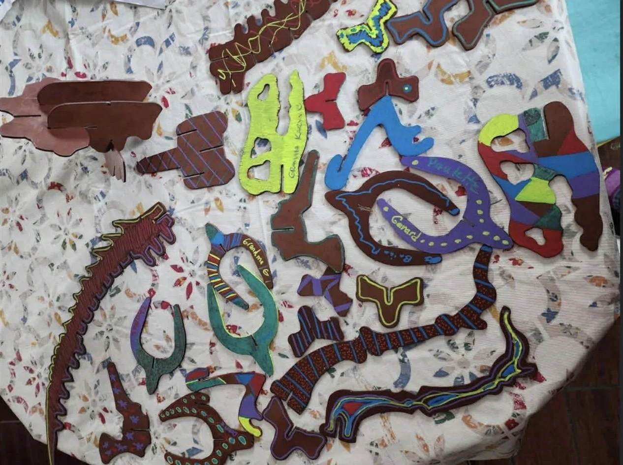 Colorful handmade cutouts of dinosaurs decorated with various patterns, words, and names, arranged on a white tablecloth with floral patterns.