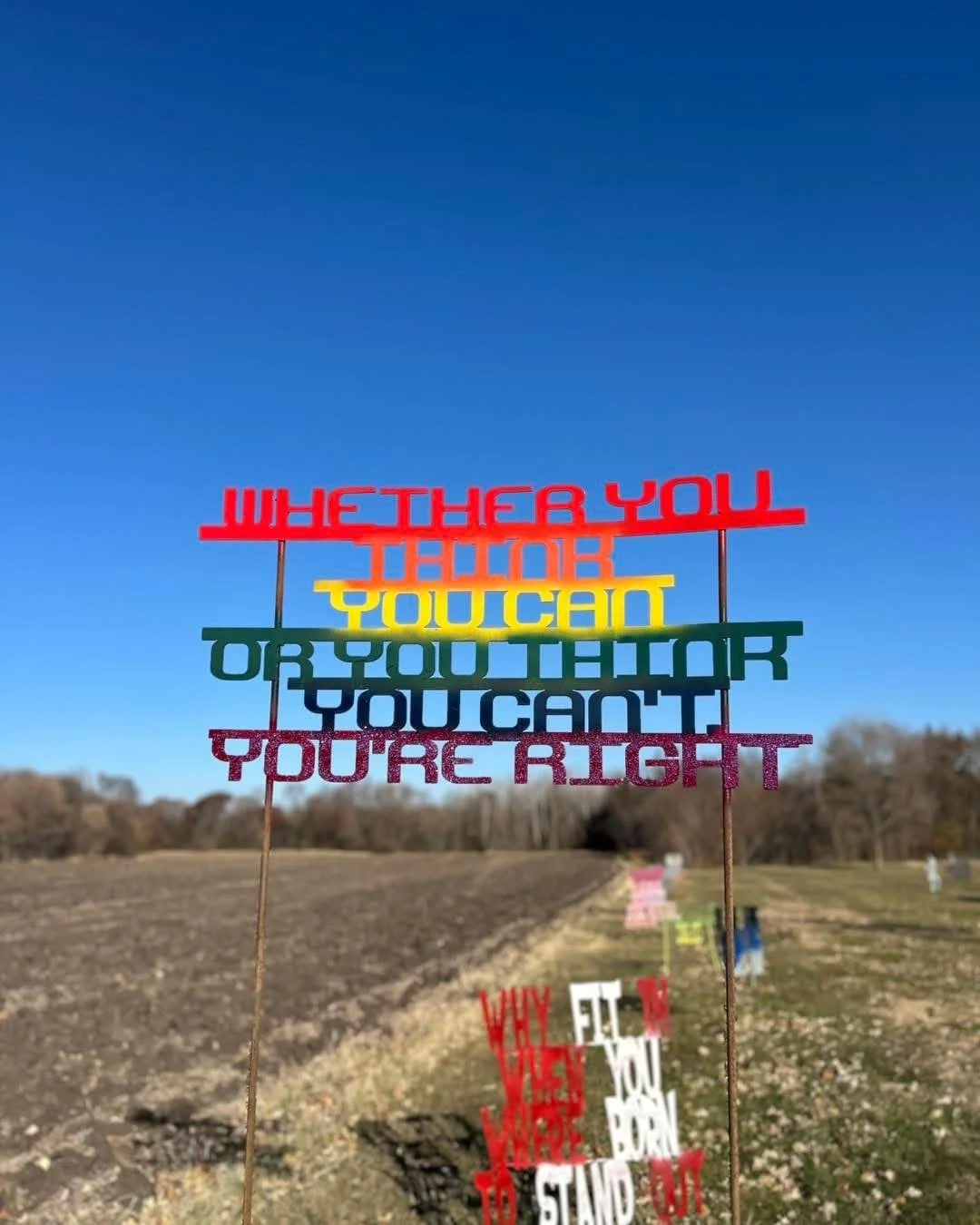 Colorful sign with motivational message on sticks in an outdoor field under a blue sky.