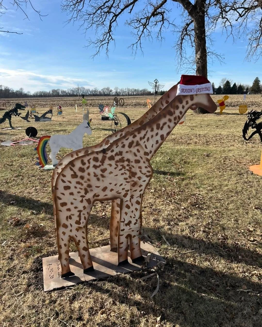 Decorative metal sculpture of a giraffe wearing a Santa hat with a "Season's Greetings" banner, set in a field with various animal and fantastical sculptures in the background on a clear day.