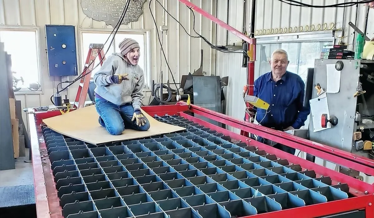 Woman kneeling on a large metal grid frame, holding a tool, with an excited expression, and an older man standing next to her in a workshop setting.