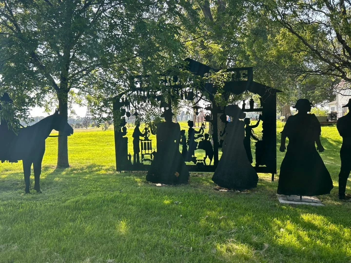 Silhouette of life-sized metal cutouts of people, a horse, and a dog in a park, with trees and grass in the background.