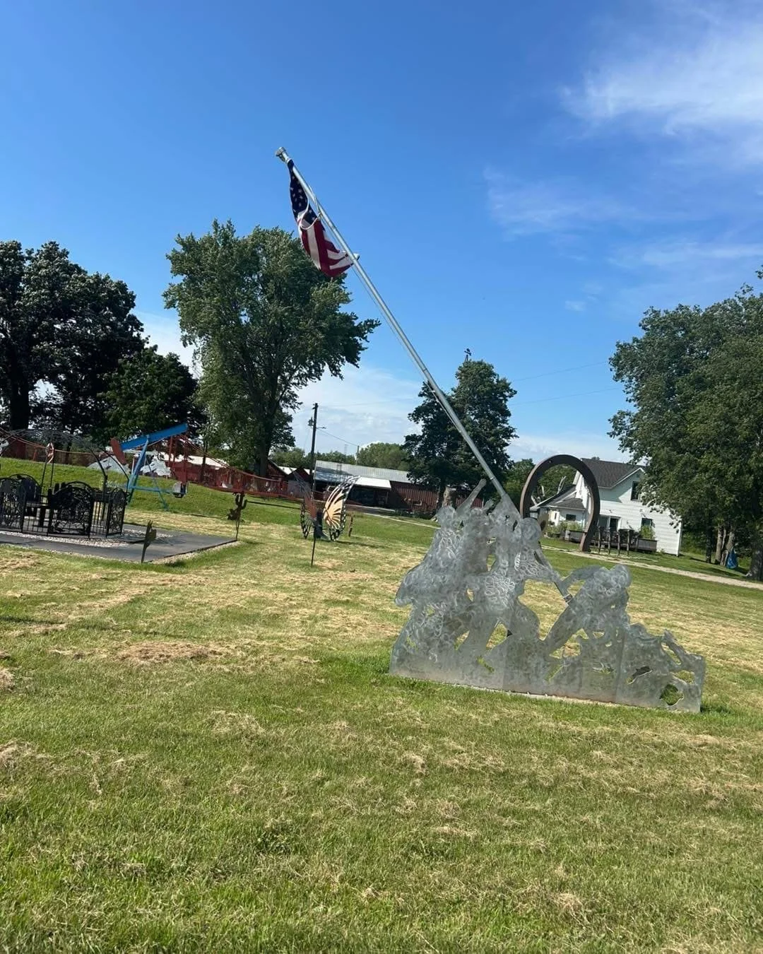 An outdoor park with a metal sculpture of cowboys on horseback, an American flag on a tall pole, and various playground equipment in the background, under a partly cloudy sky.