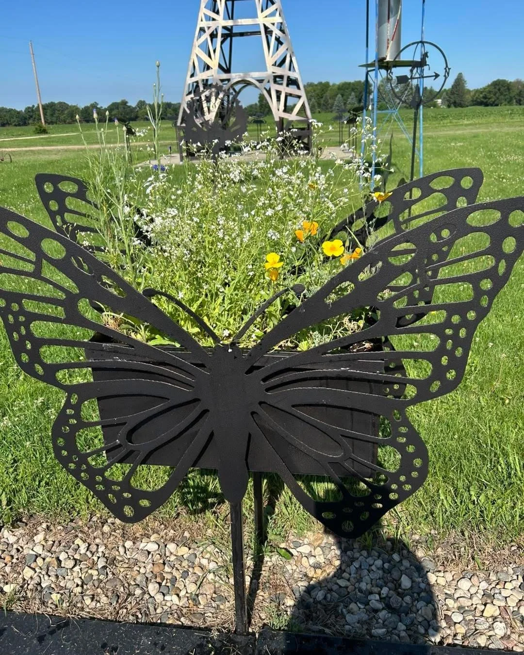 Black butterfly-shaped metal flower planter with yellow and white flowers, set outdoors on a grassy area with a windmill and other metal structures in the background.