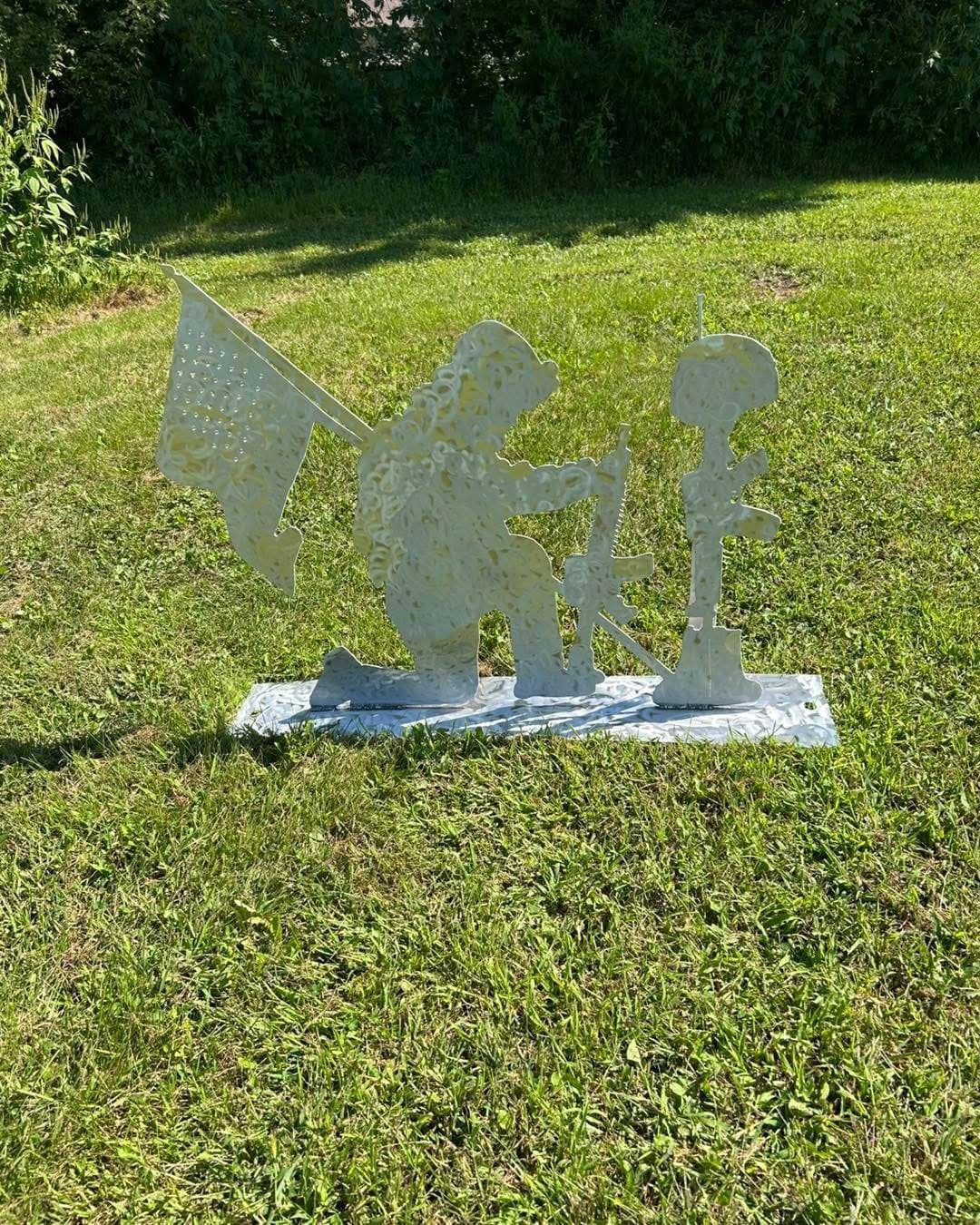 Metal garden sculpture of a person kneeling and working with a small robot or camera on a tripod, outdoors on grass.