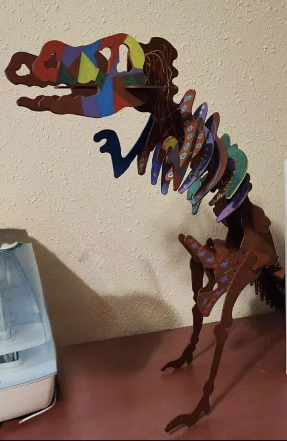 Colorful wooden sculpture of a dinosaur, possibly a T-Rex, with a rainbow-colored head, neck, and back, standing on a reddish surface near a light-colored wall.
