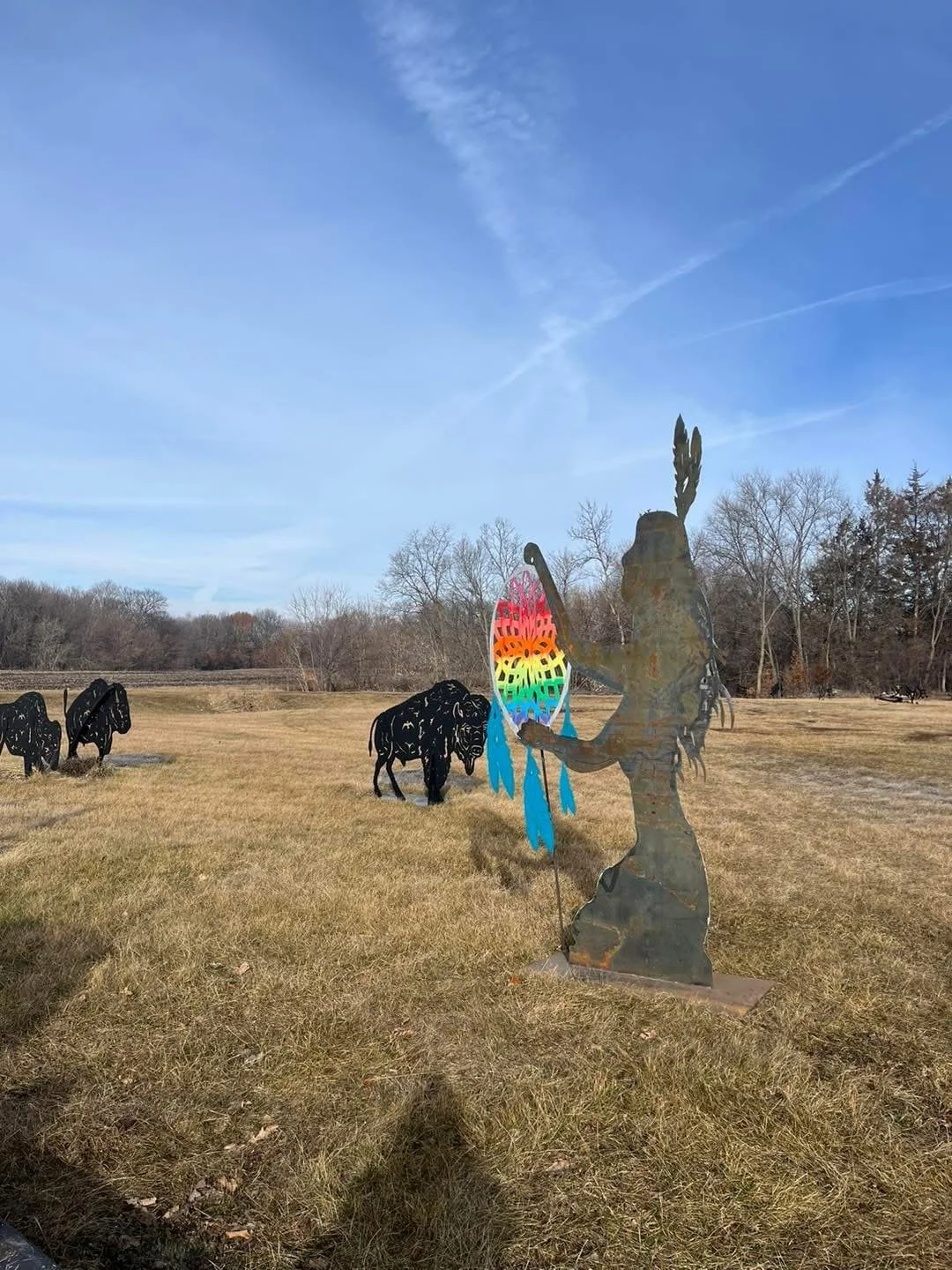Open field with leafless trees, metal sculpture of a woman with a feather in her hair holding a colorful dream catcher, and black metal bison in the background against a bright blue sky with wispy clouds.